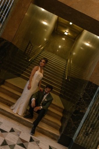 A woman in a white dress and a man in a suit pose on stairs in a building with marble walls and golden lighting.