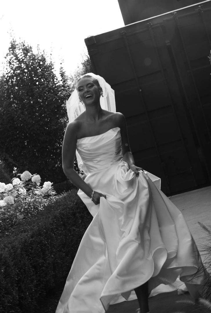 Woman in wedding dress smiling outdoors, holding her dress, with trees and a structure in the background.