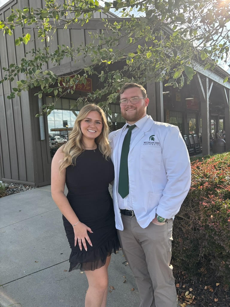 A smiling woman in a black dress and a man in a white shirt and green tie stand outdoors in front of a building with large windows and greenery.