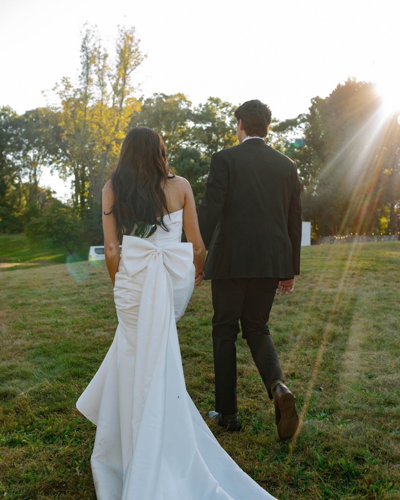 A couple in formal attire walking on grass with trees and sunlight in the background.