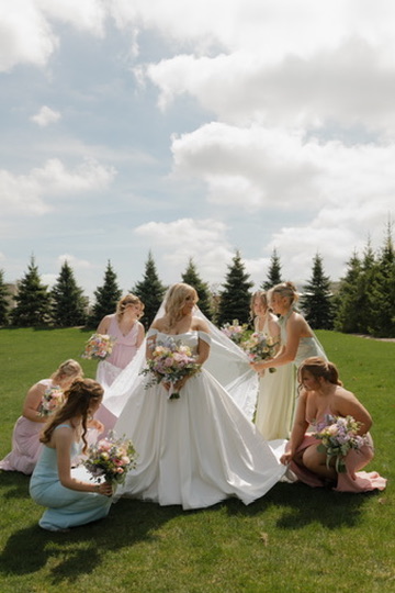 Bride in white gown surrounded by five women holding bouquets outdoors on grass, with trees and cloudy sky in background.