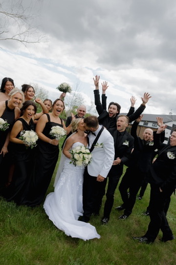 Group of wedding guests and a bride and groom outdoors, celebrating and smiling, with cloudy sky in background.