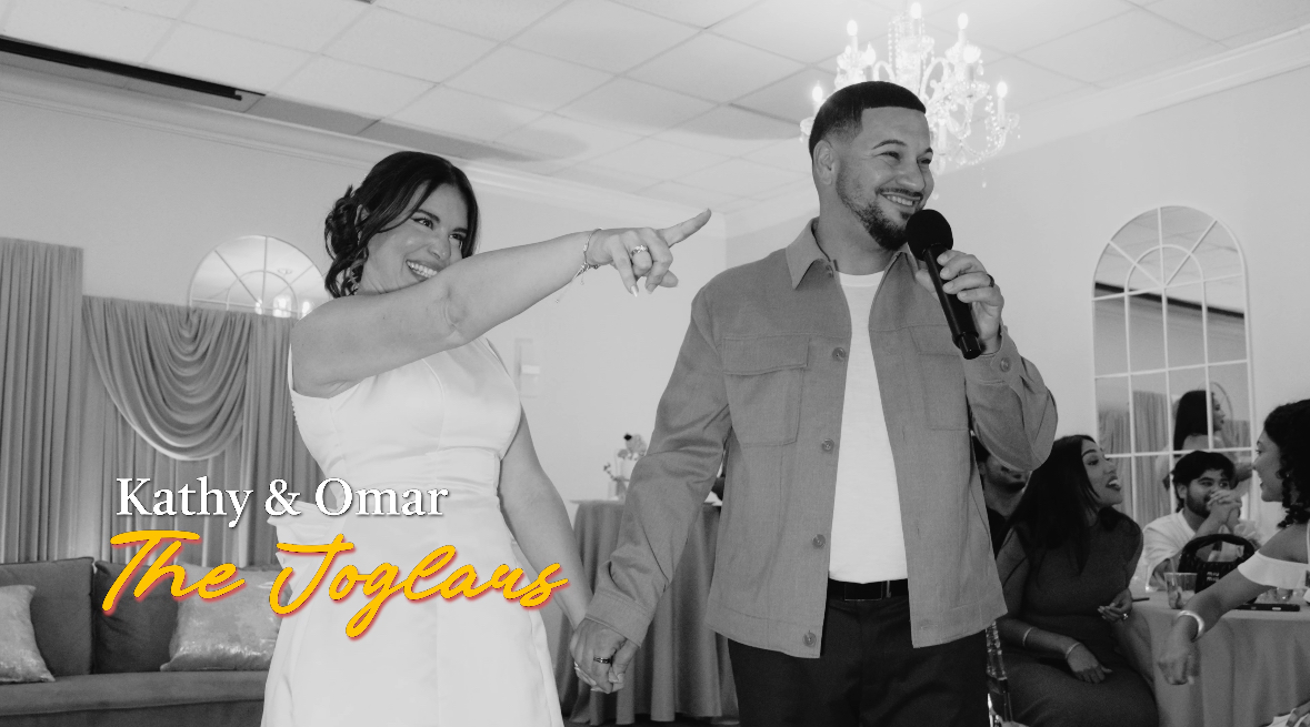 Kathy and Omar at a celebration, with Kathy pointing and Omar holding a microphone, in a decorated indoor venue.