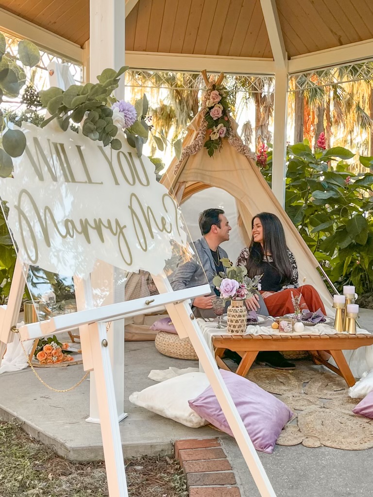 Couple sitting on a low table inside a decorated outdoor gazebo, with a sign reading 'Will You Marry Me' reflected in the glass.