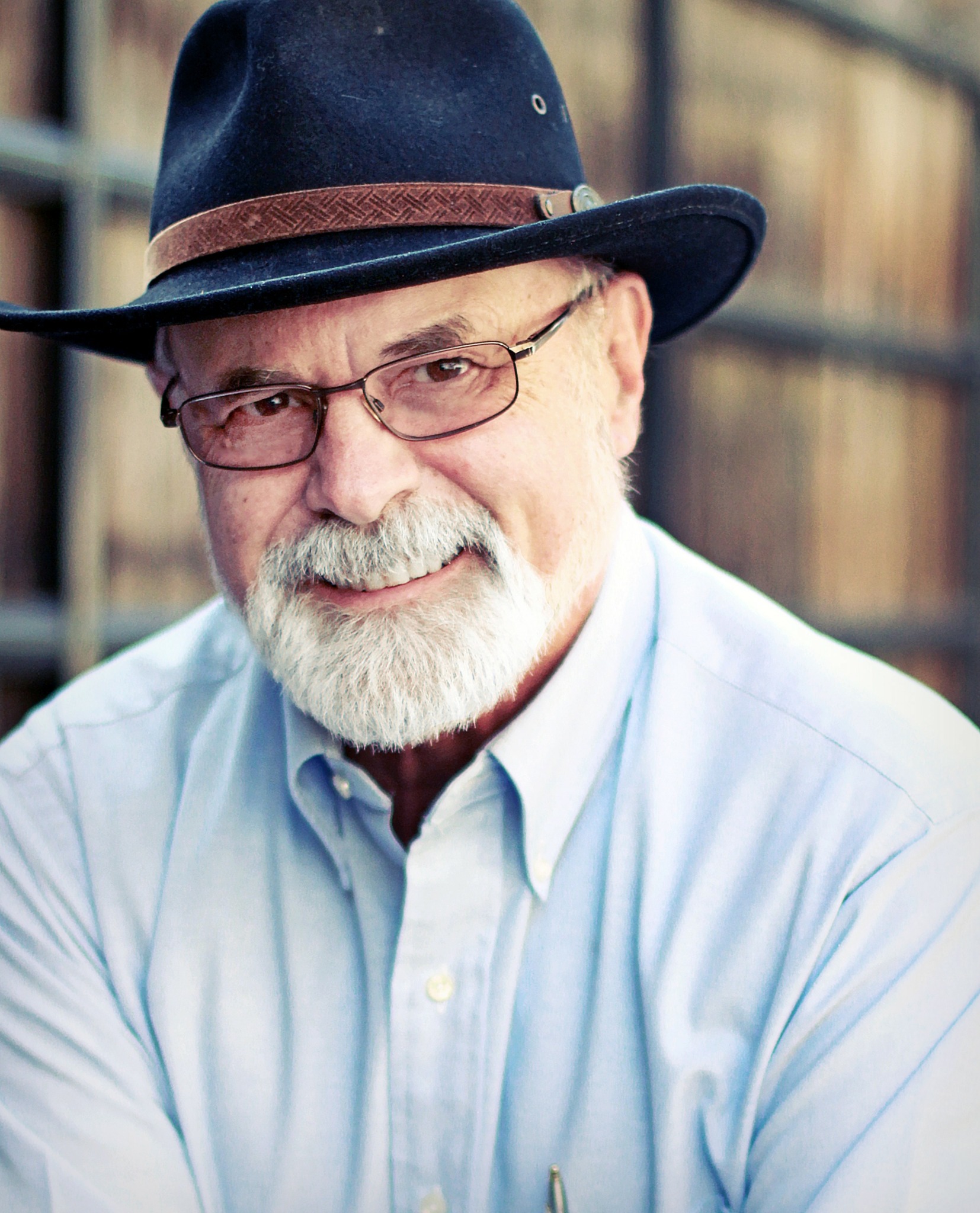 Older man with glasses, white beard, wearing a hat and light blue shirt, smiling outdoors.