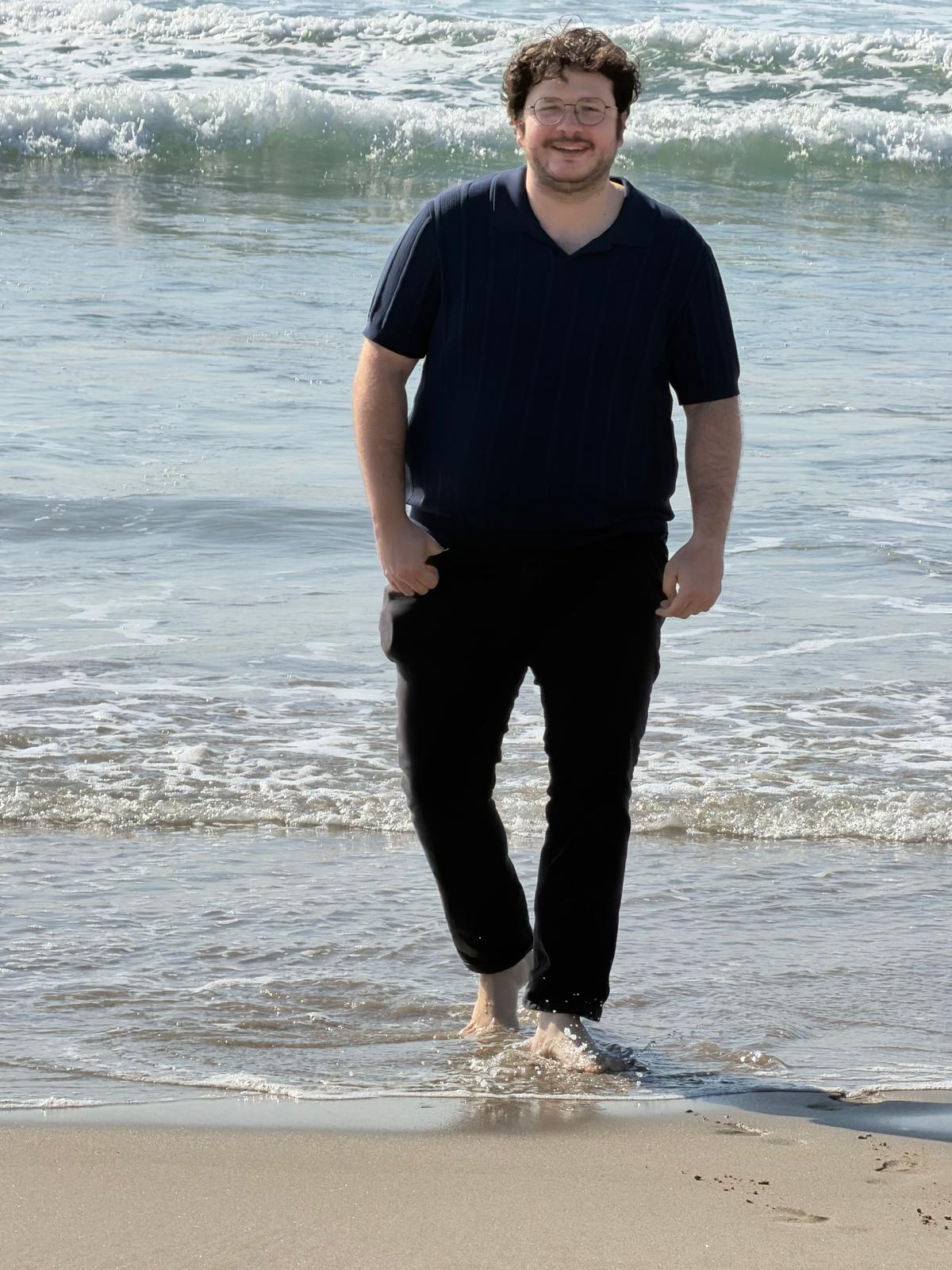 Person walking in shallow ocean water at the beach, waves in background, smiling, wearing dark clothing.