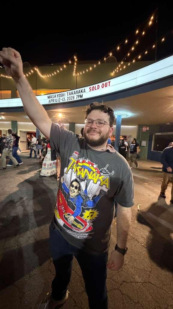 Man with glasses and beard smiling, raising fist, wearing a Tijuana-themed T-shirt, in a busy outdoor area at night.