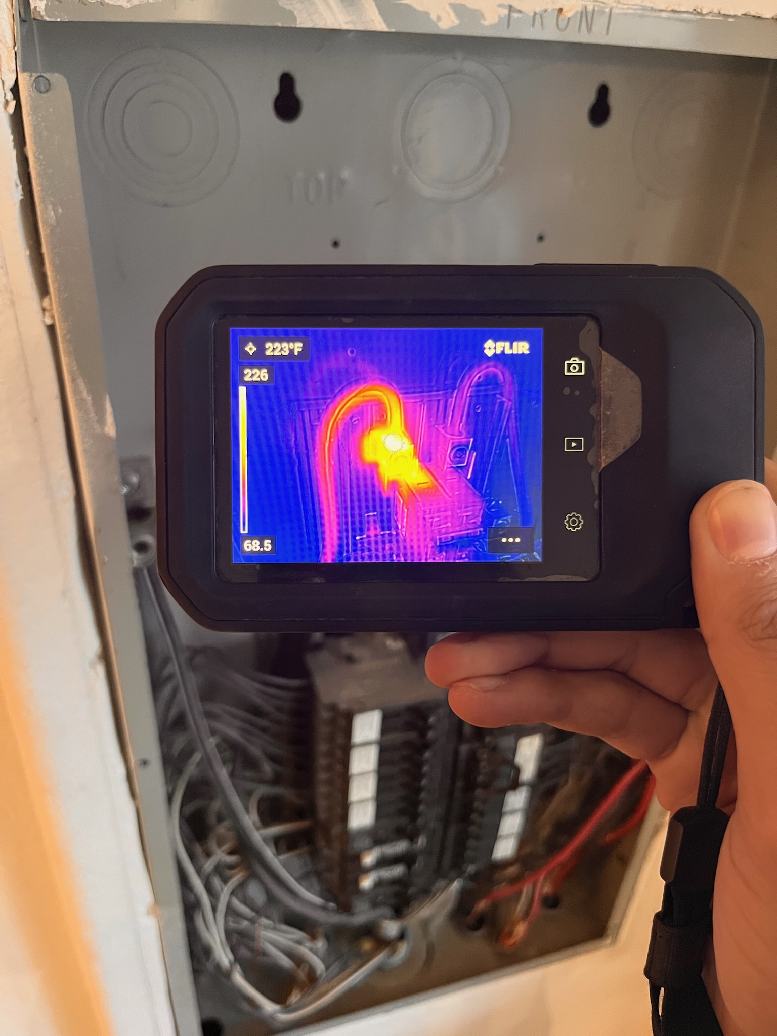 Person holding a thermal camera showing a colorful thermal image of an electrical panel with wires inside.