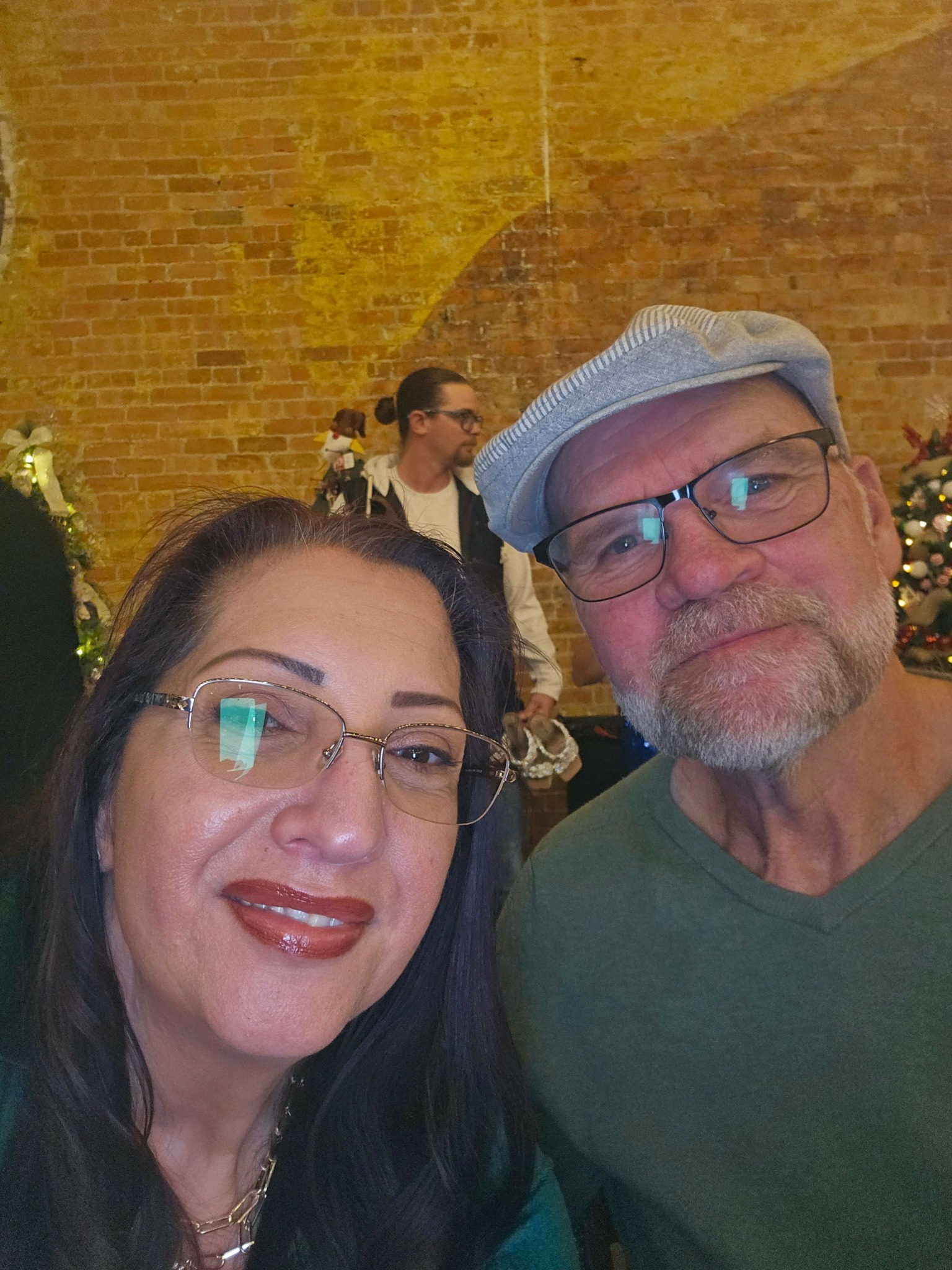 Two people smiling, one woman with glasses and long dark hair, and one man with glasses and a cap, in front of a brick wall.
