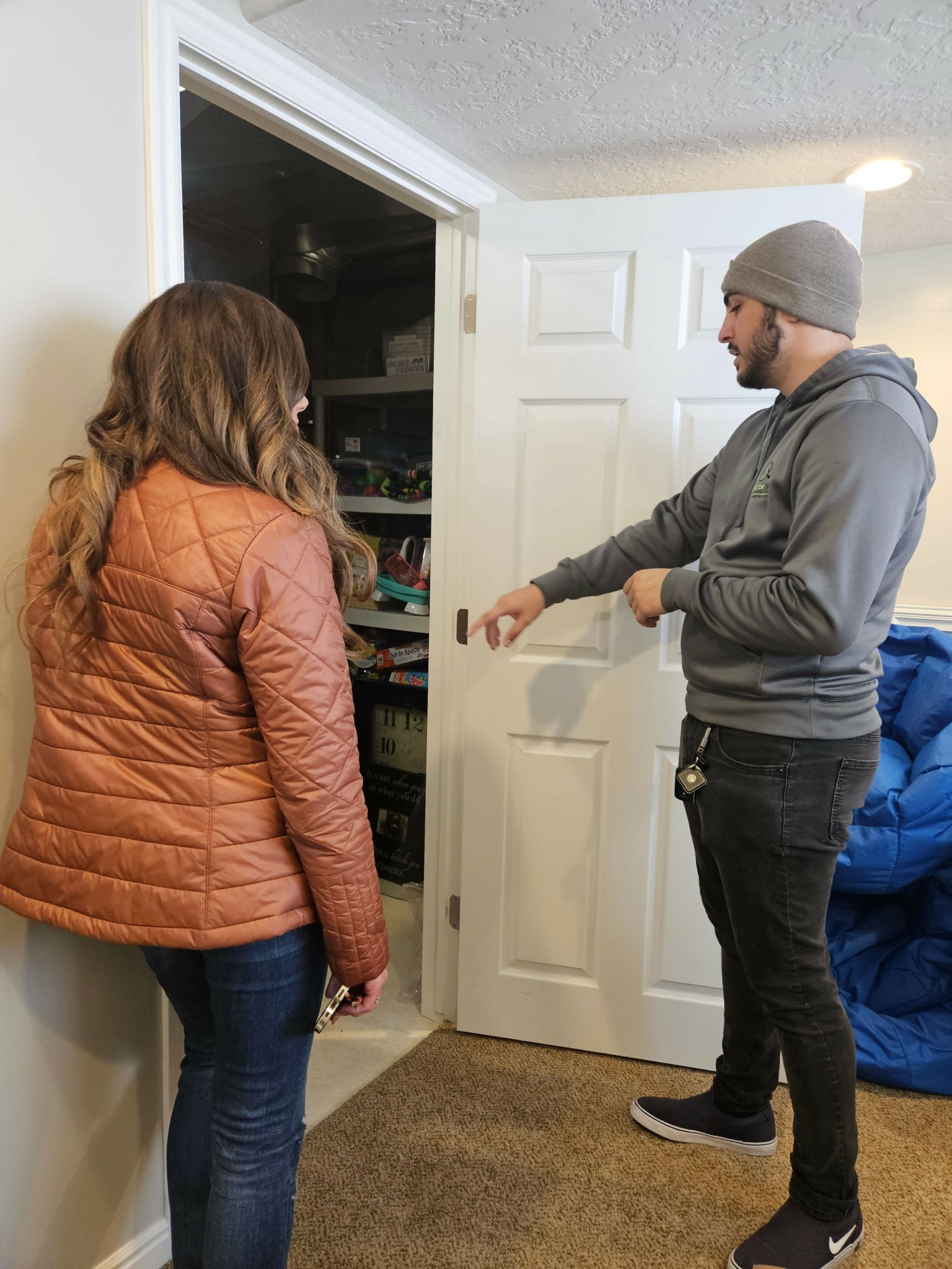 Two people standing indoors, one wearing a gray hoodie and beanie, the other in an orange jacket, facing each other.