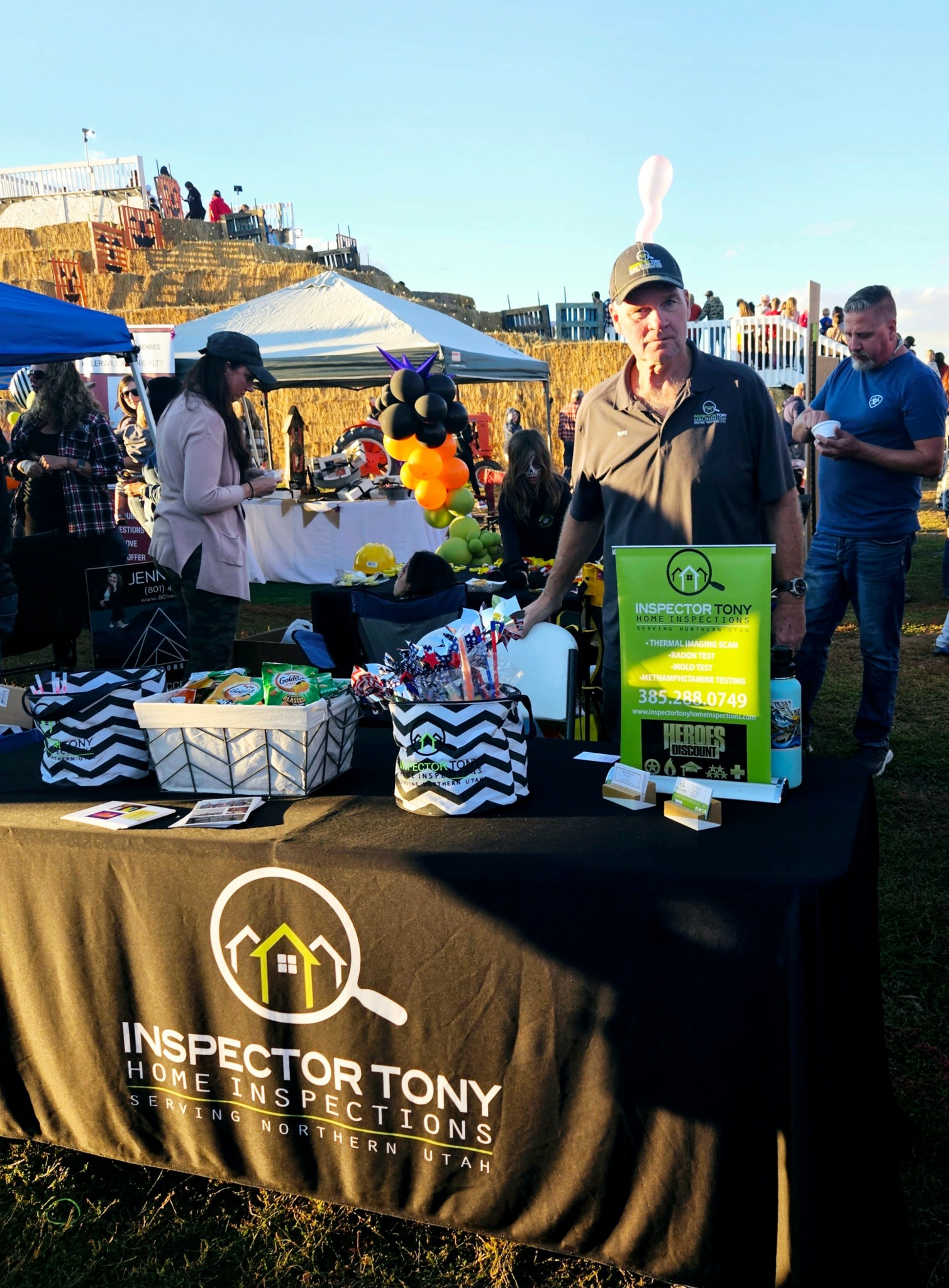 Man wearing sunglasses and a cap stands behind a table with items, outdoors with tents and people in background.