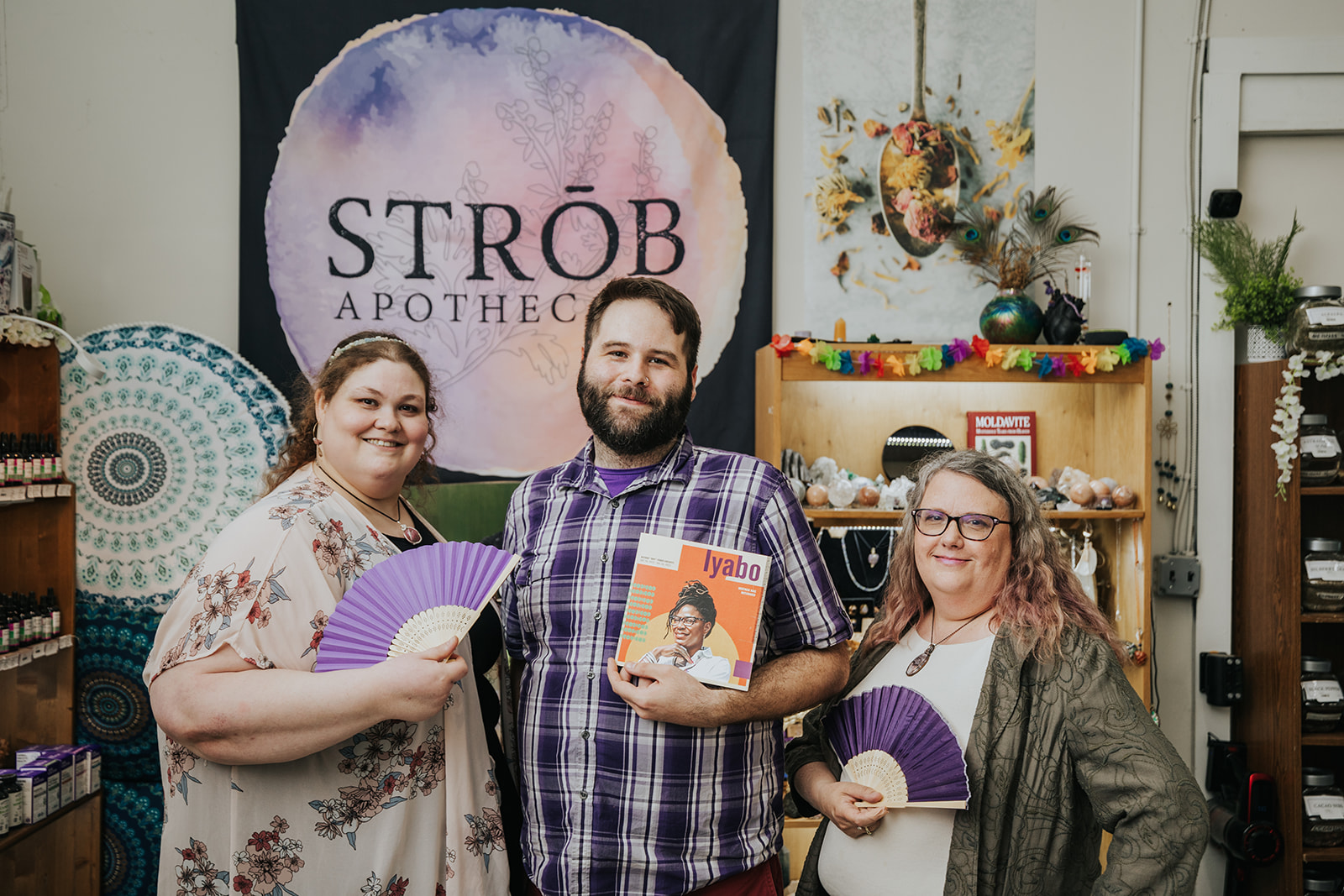 Three people standing indoors, holding fans and a book, with a banner reading 'STROBE APOTHEC' in the background.