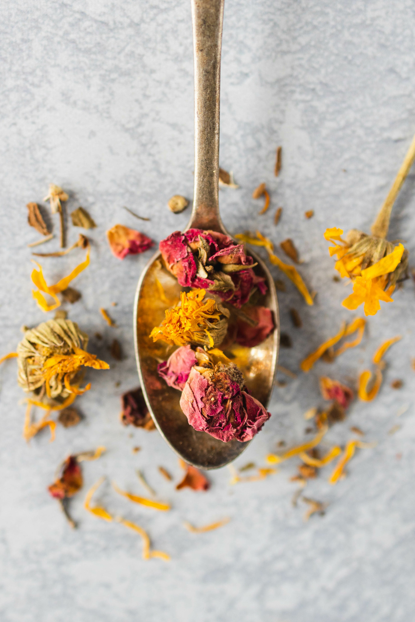 Spoon with dried flower petals and herbs, scattered on a light surface.