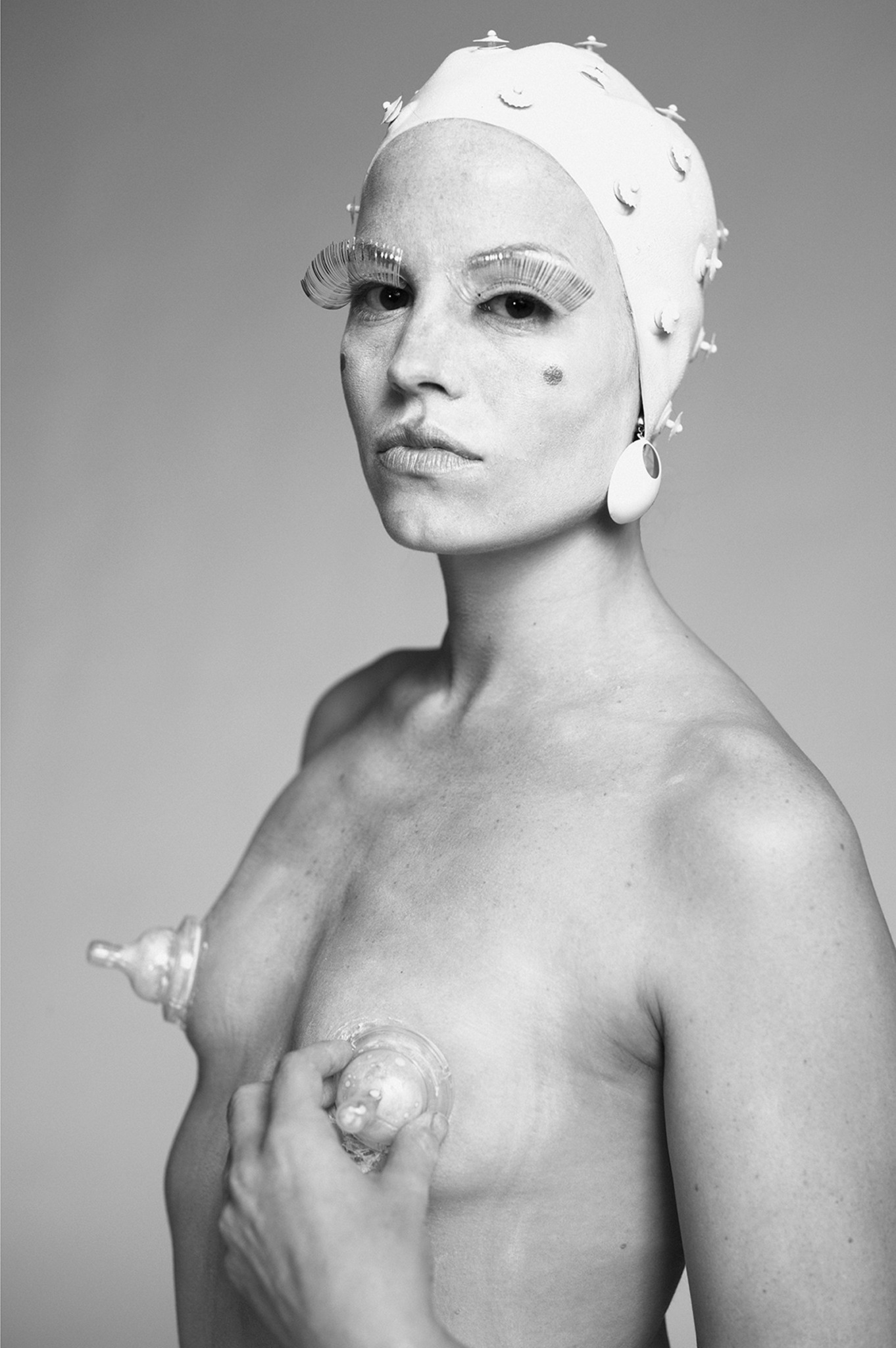Black and white portrait of a topless woman with a head covering and large earrings, holding a seashell to her chest.