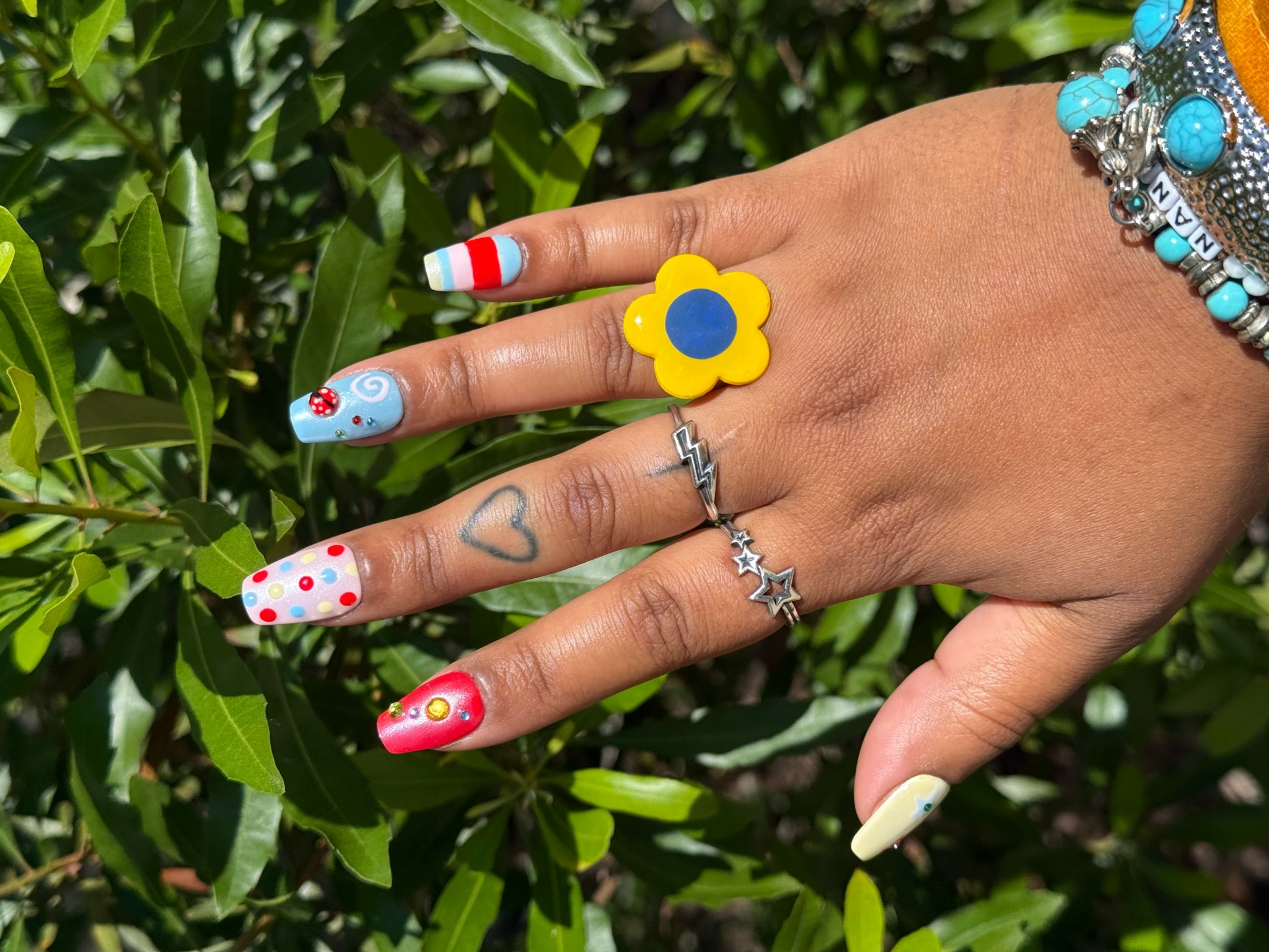 Hand with colorful nail art, rings, and bracelets against green leaves background.