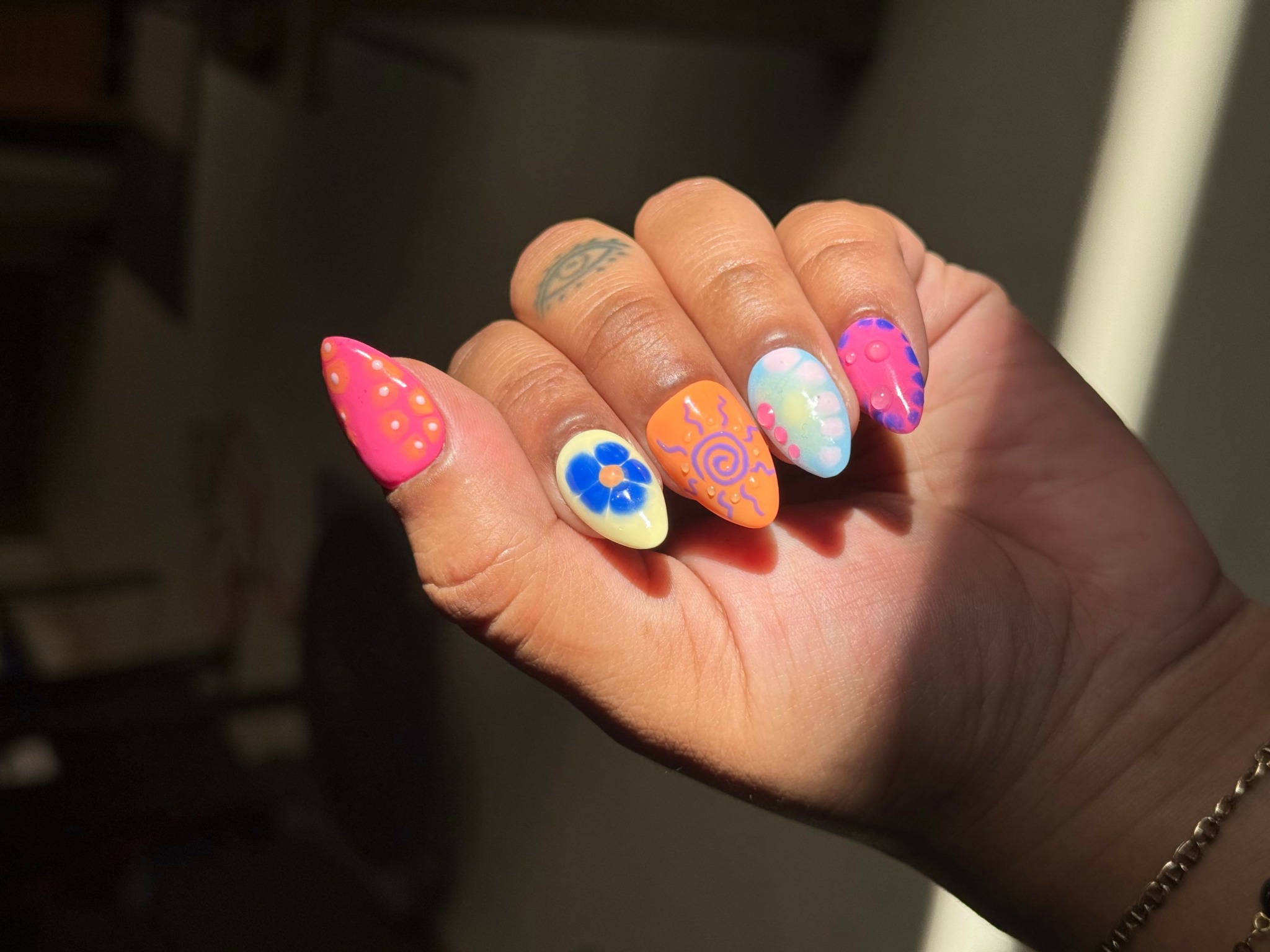Hand with colorful, decorated nails featuring pink, blue, orange, and white designs, including flowers and swirls, in natural lighting.