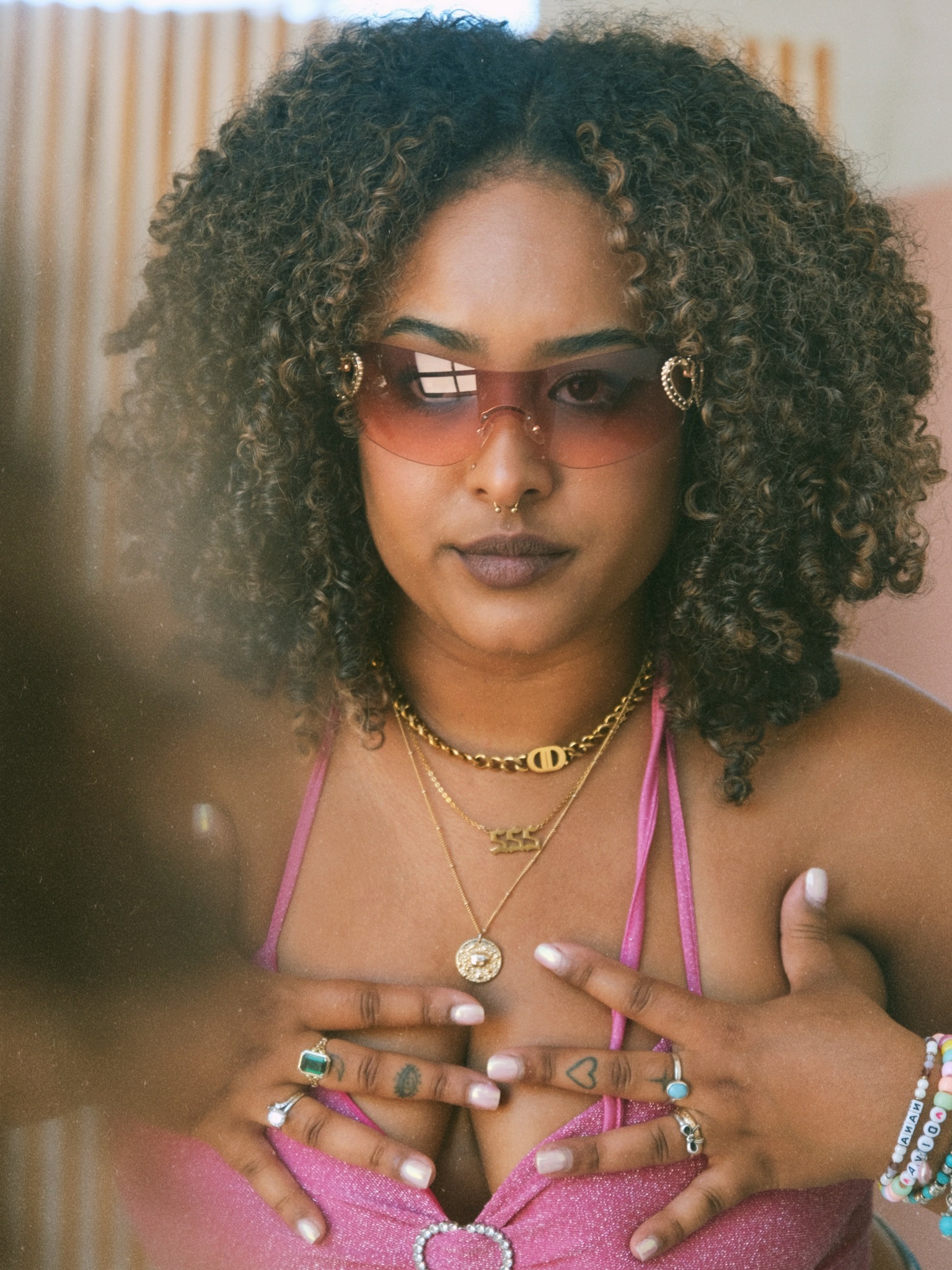 Woman with curly hair wearing sunglasses, layered necklaces, and a pink top, holding her chest with hands adorned with rings and bracelets.