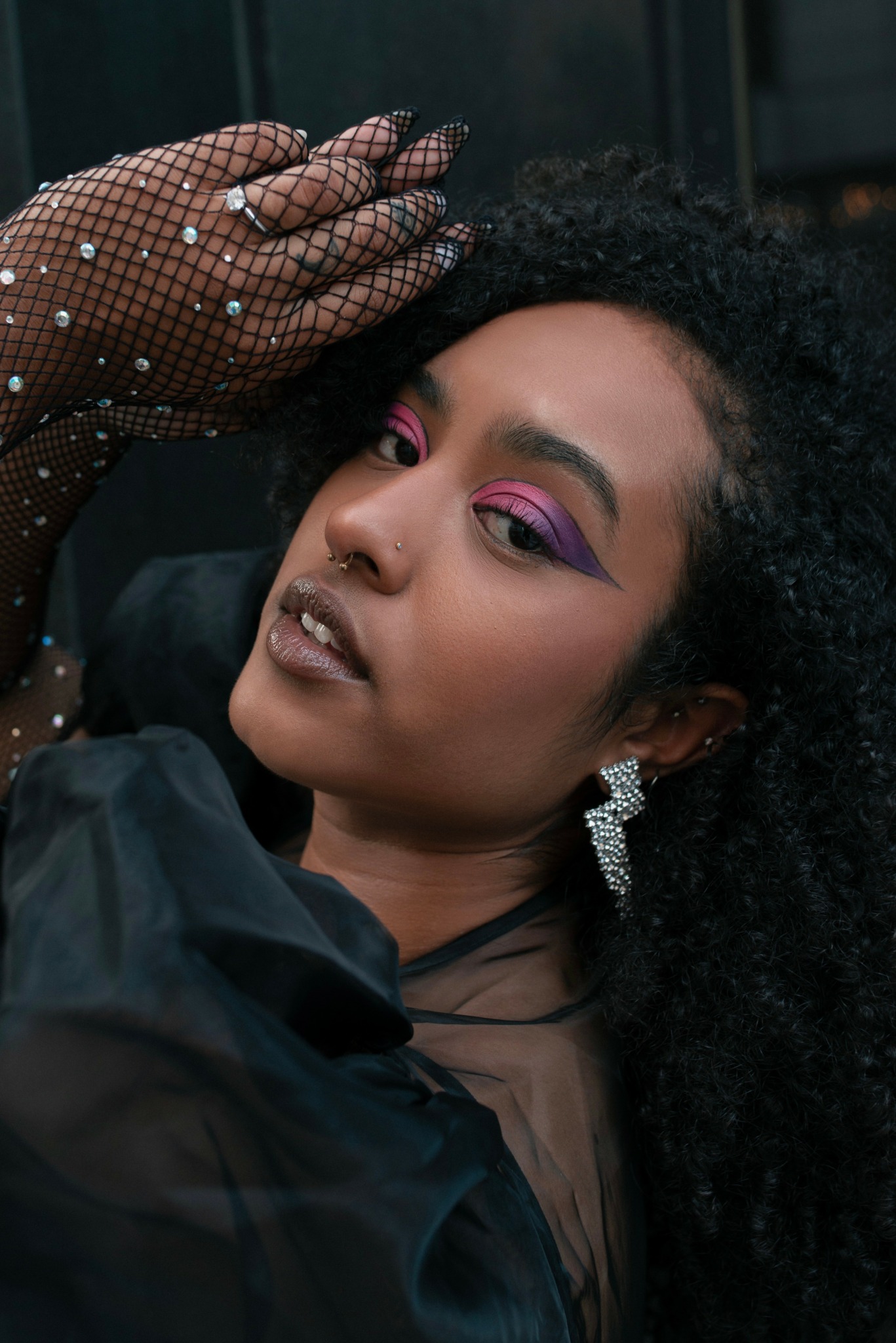 Close-up of a woman with curly hair, vibrant makeup, and jewelry, posing with her hand on her head.