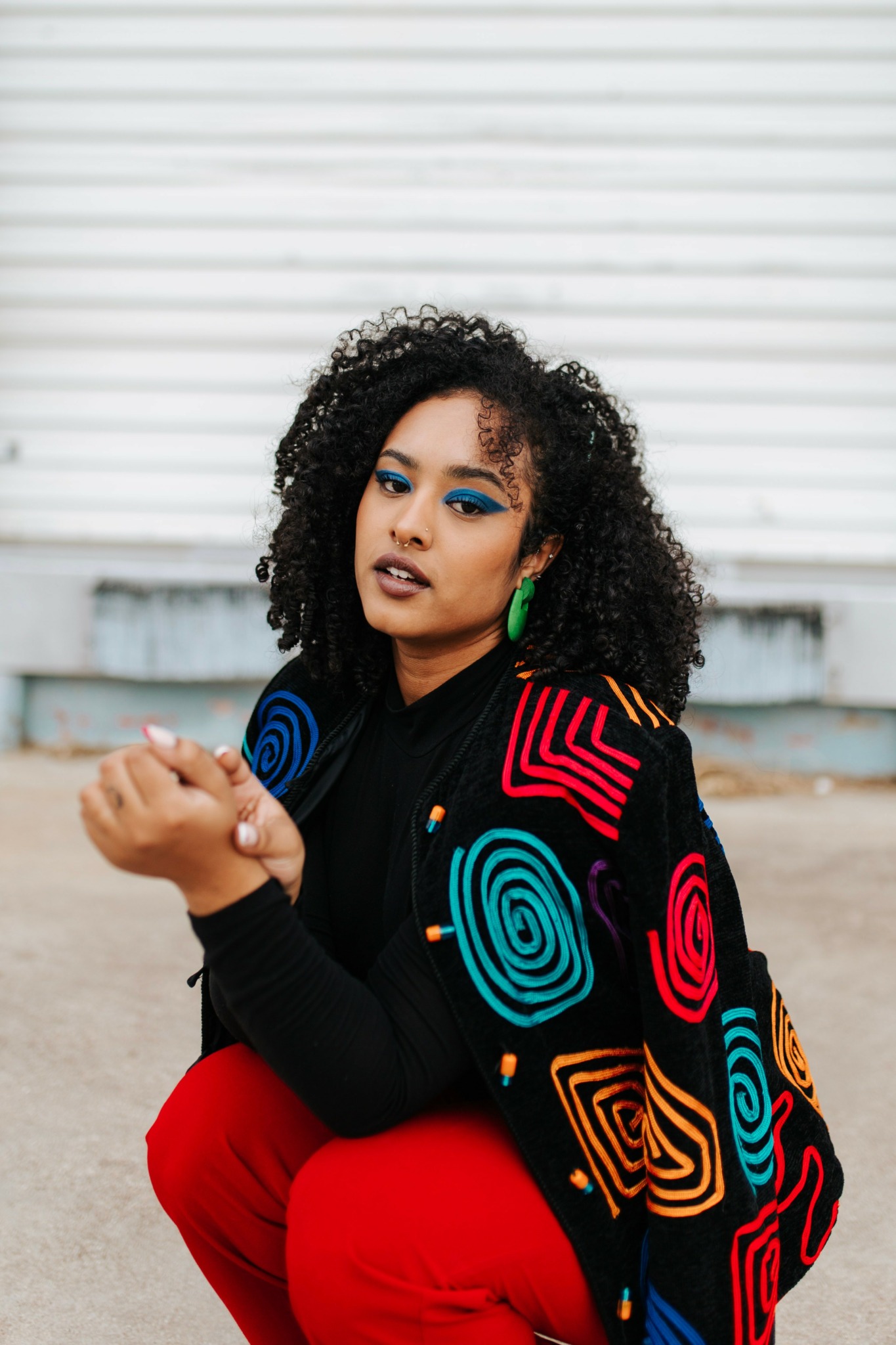 Young woman with curly hair, wearing colorful makeup and a patterned jacket, sitting outdoors against a white background.