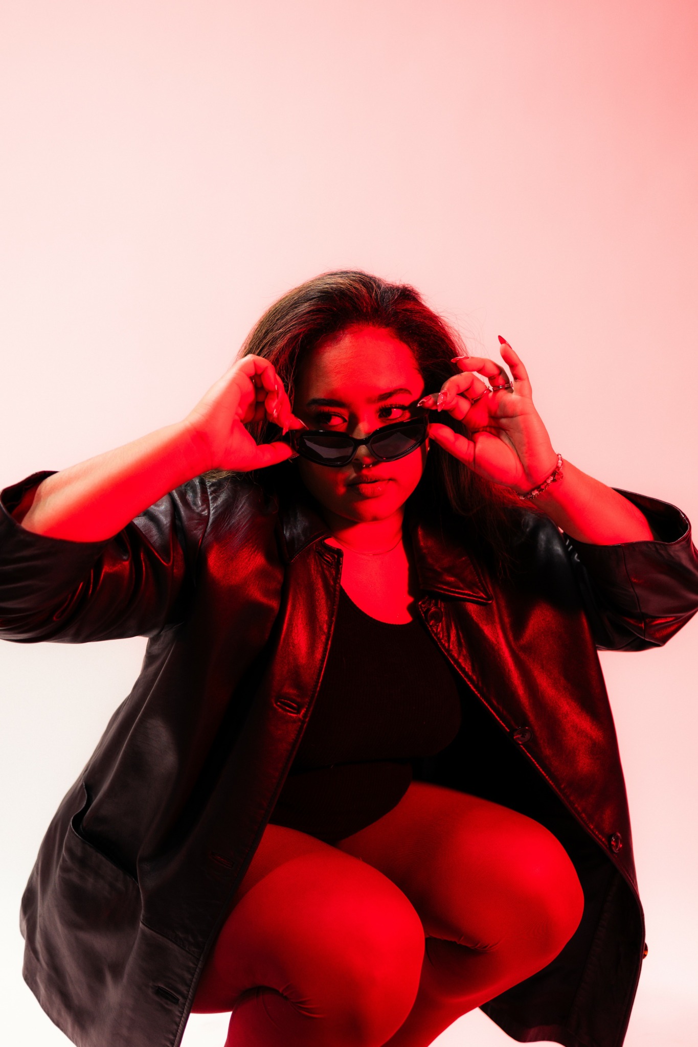 Person with long hair wearing sunglasses, black jacket, and black top, squatting against a pink background.