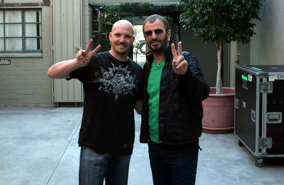 Two men standing outdoors, smiling and making peace signs, with trees and a building in the background.