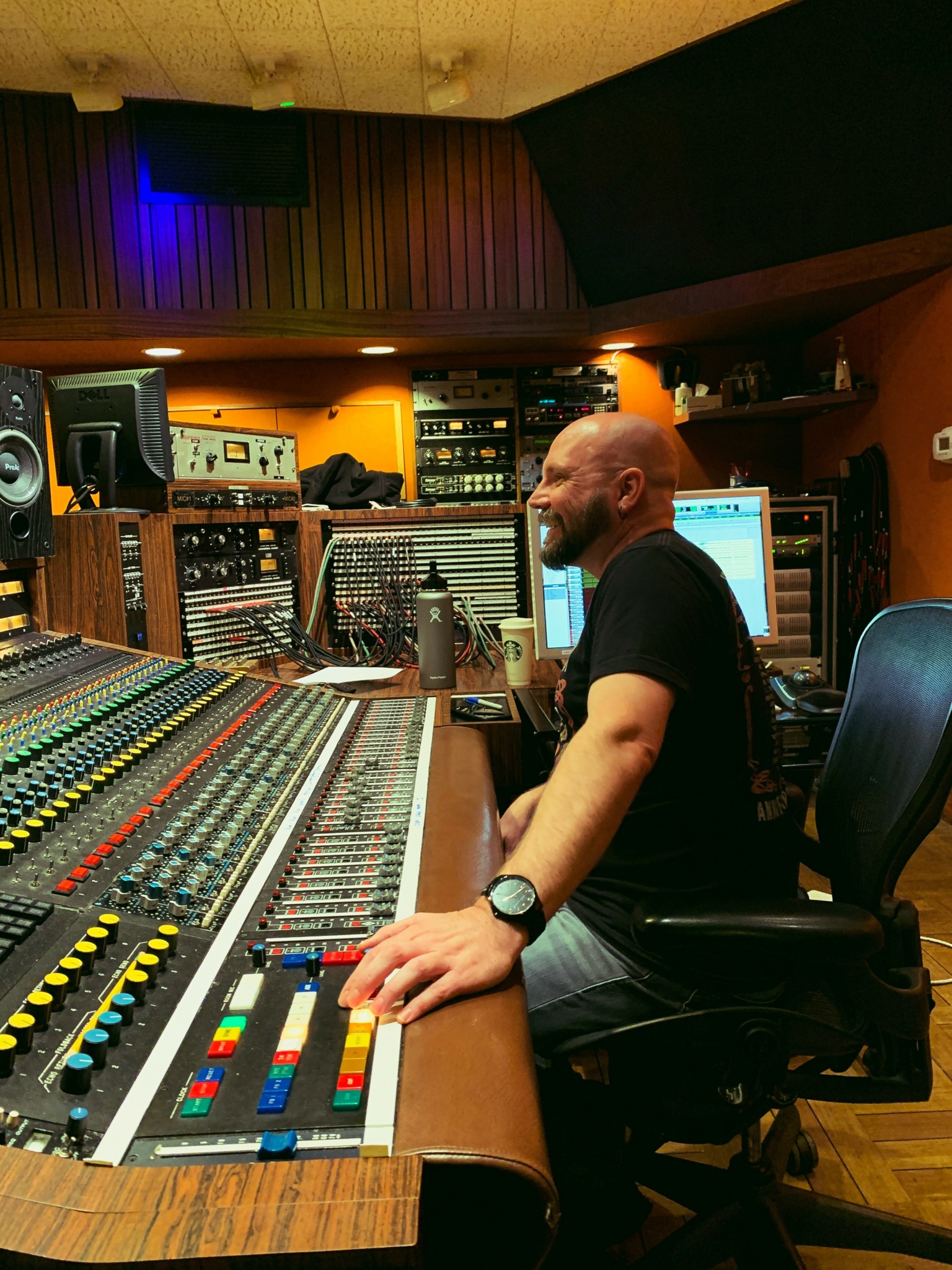 Bald man in black shirt sitting at mixing console in recording studio with equipment and computer screens.