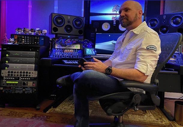 Man sitting in a music studio with audio equipment and speakers, holding a device, wearing a white shirt, smiling.