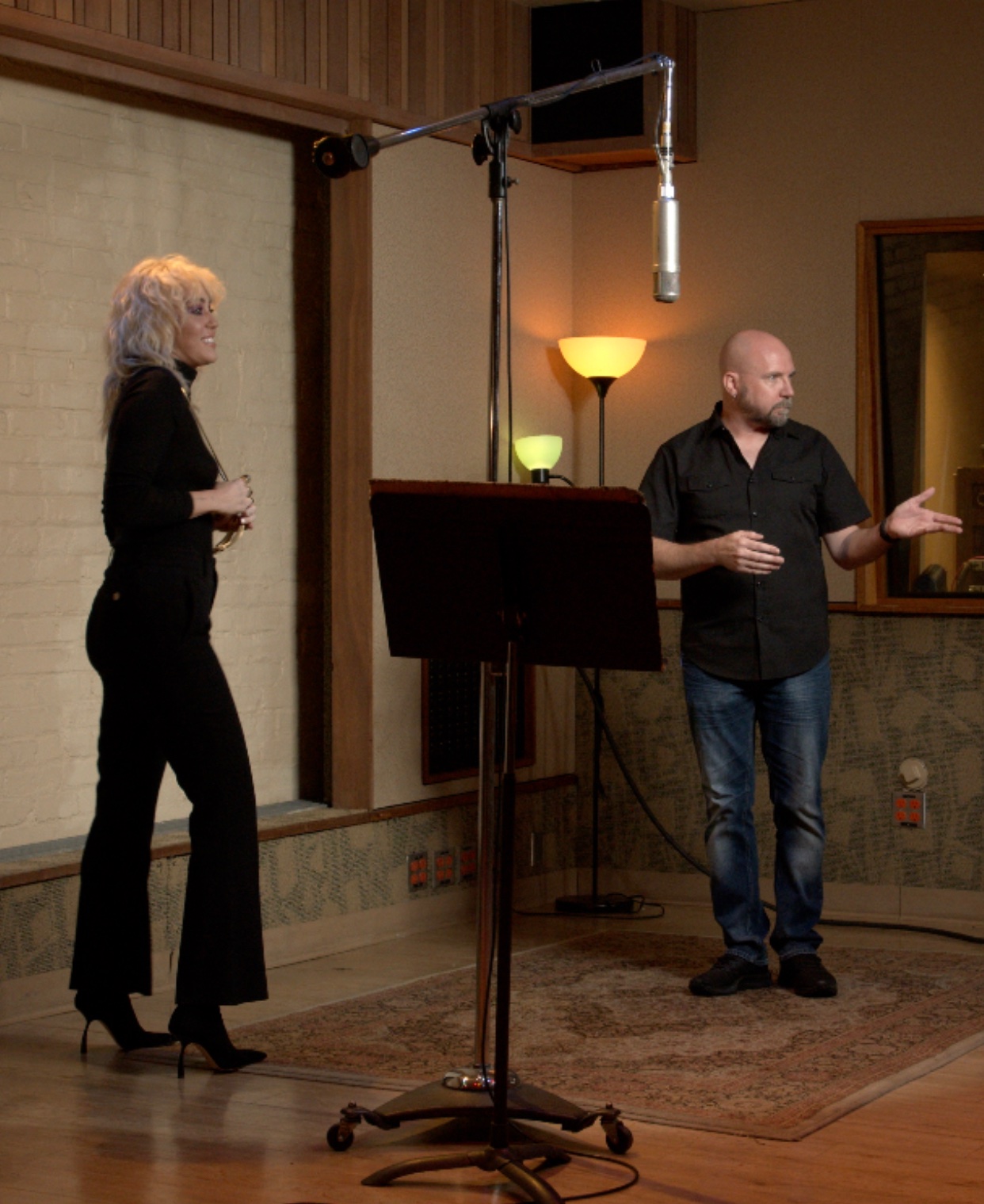 Two people in a recording studio, one woman and one man, standing and talking, with a microphone overhead, warm lighting, and a mirror.