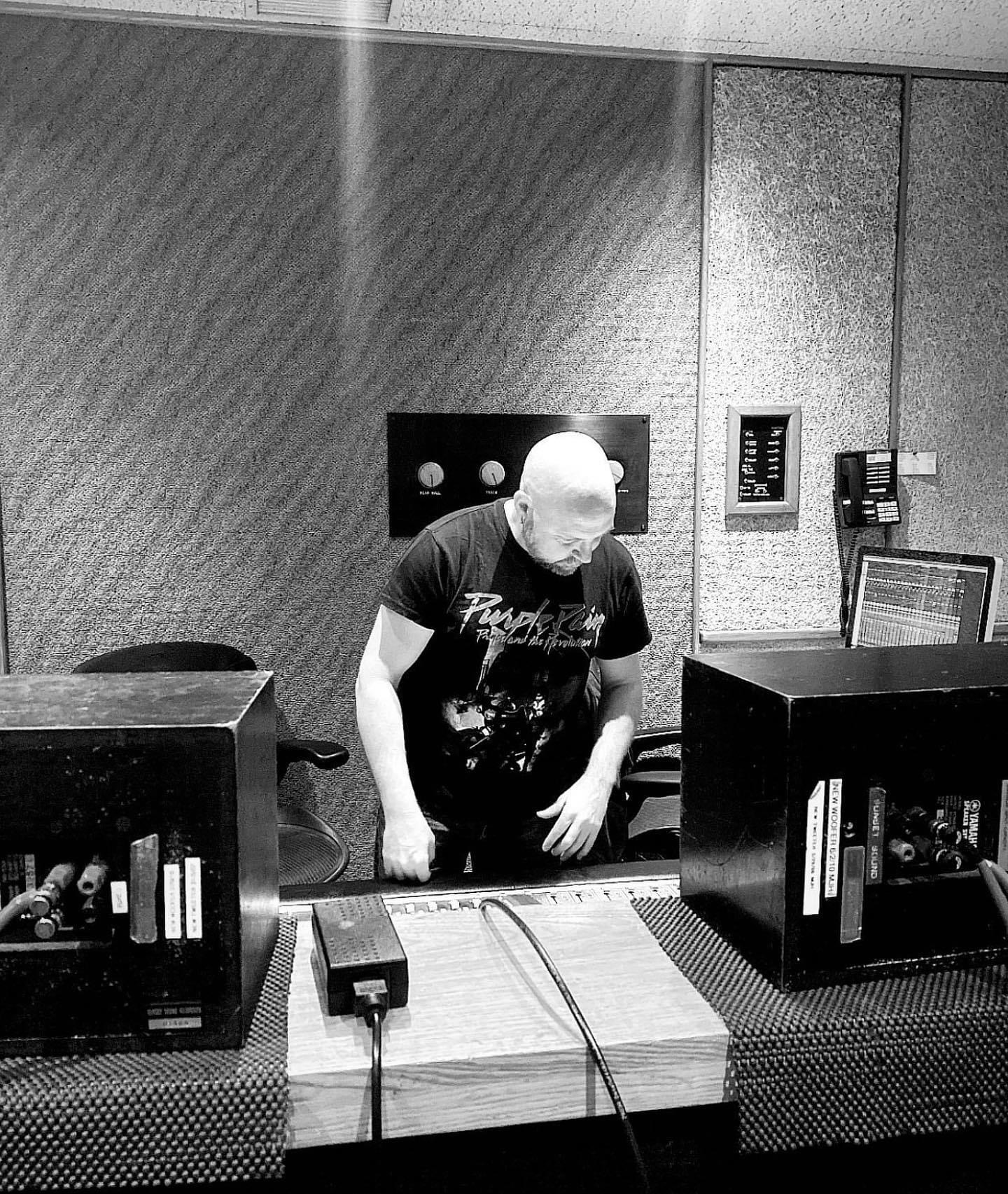 Person with a bald head wearing a black T-shirt, standing at a desk with audio equipment, in a room with textured walls.