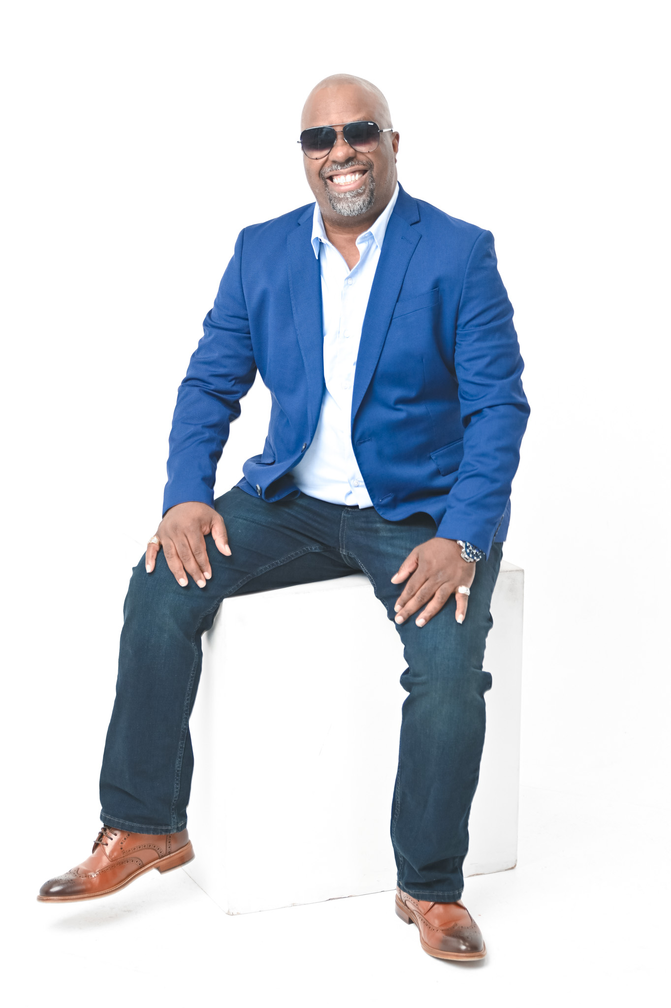Man sitting on a white block, wearing sunglasses, a blue blazer, white shirt, jeans, and brown shoes, smiling.