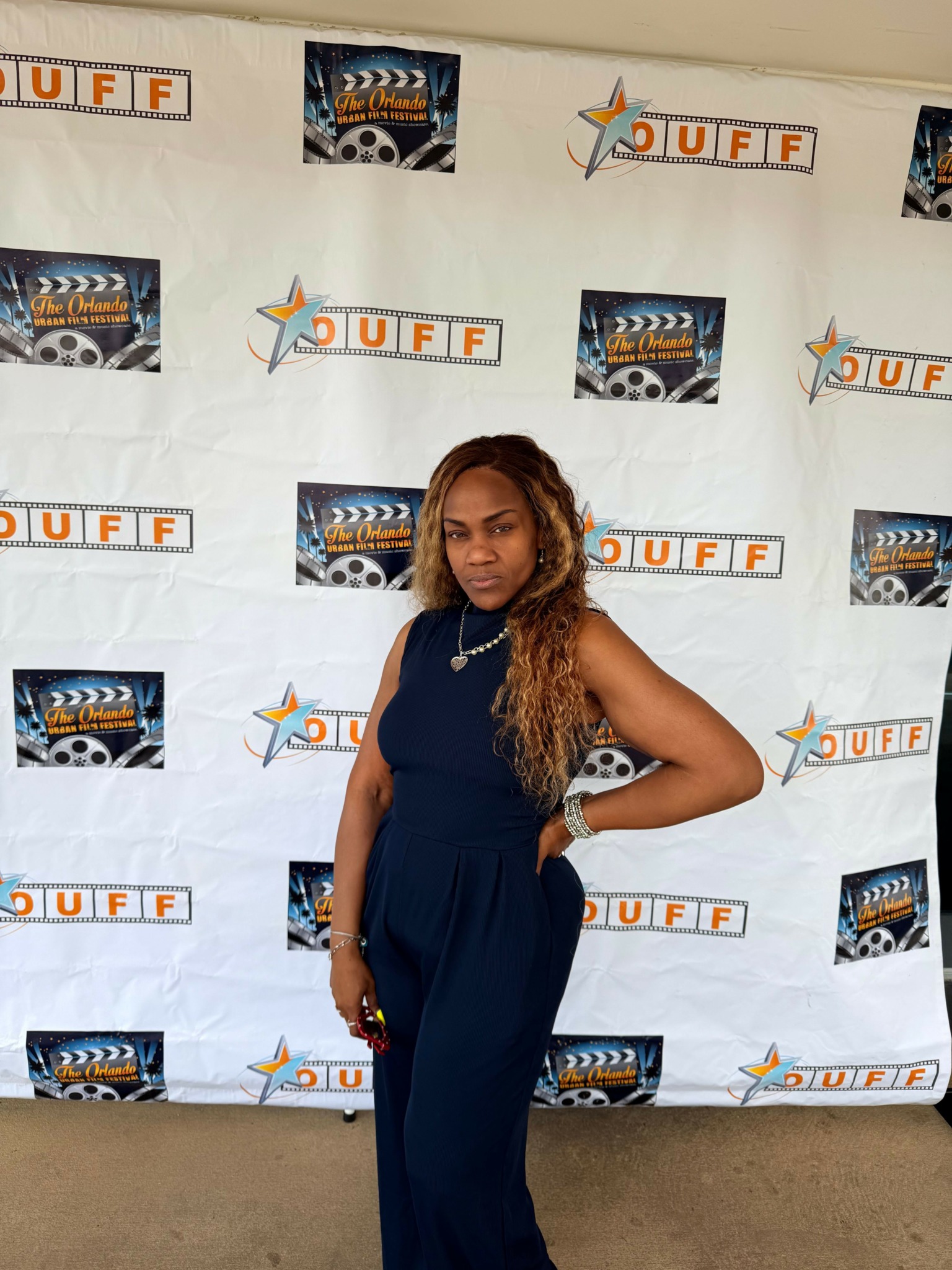 Woman with long curly hair in a dark blue dress standing in front of a backdrop with logos and text.