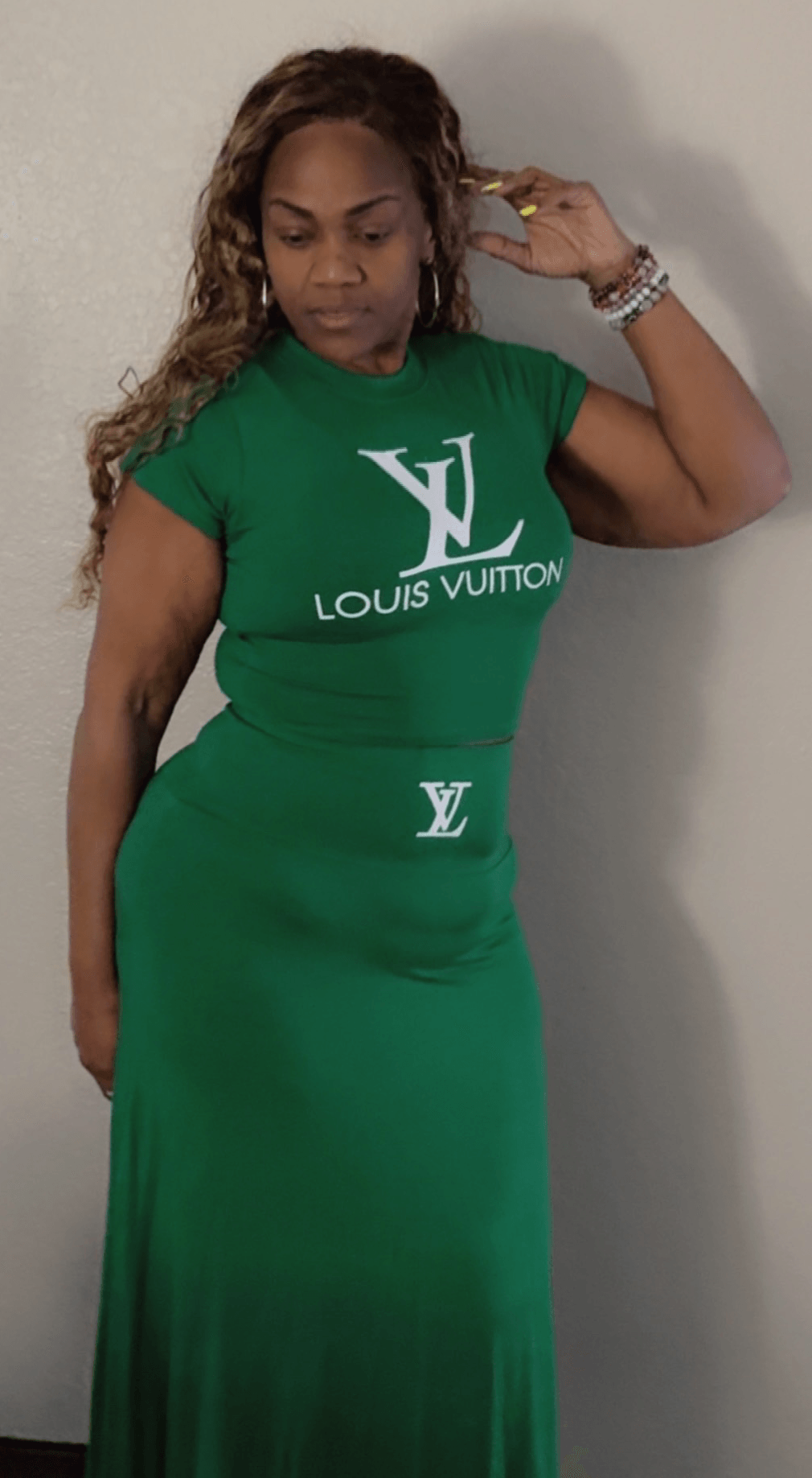 Woman in a green dress with Louis Vuitton logo, standing against a plain wall, looking down, with her hand near her head.