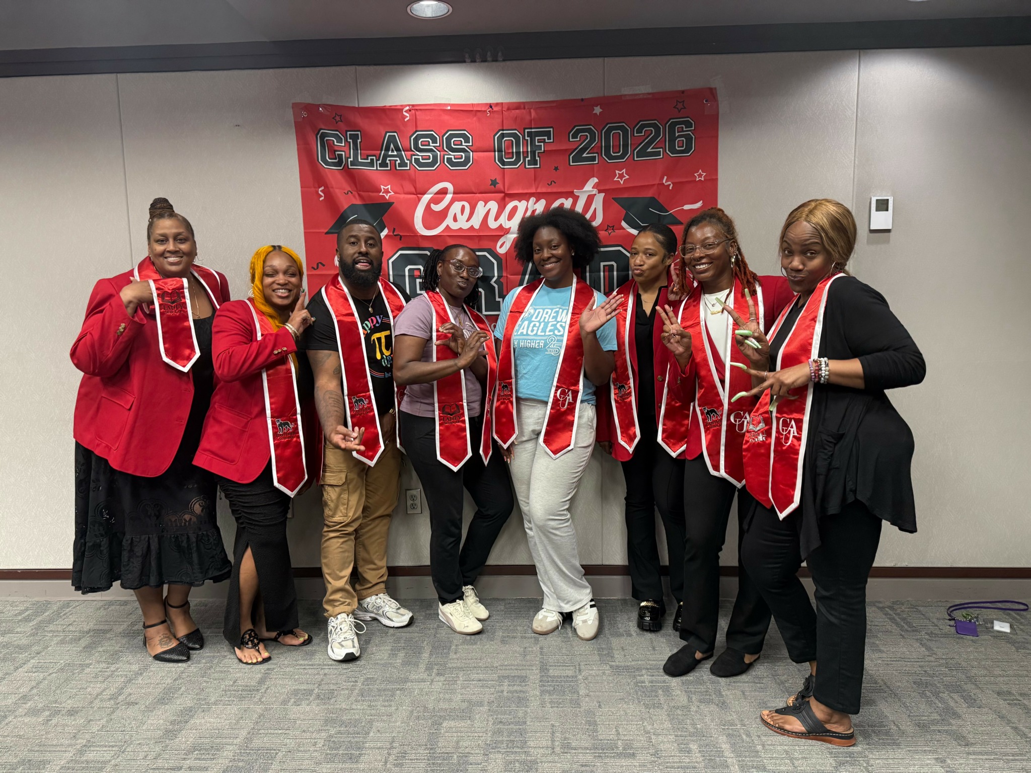 Group of eight graduates wearing red sashes and caps, standing in front of a red banner that reads 'Class of 2026 Congrat'.