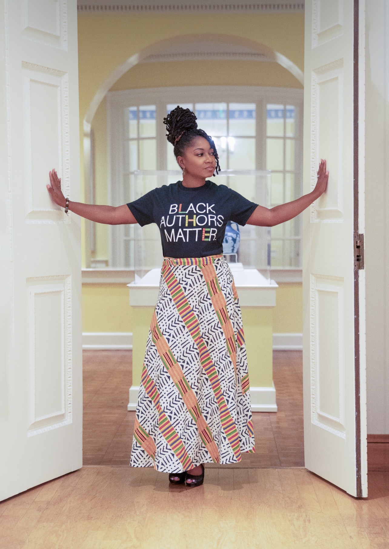 Woman standing in doorway with arms outstretched, wearing a black T-shirt and patterned skirt, in a bright room with large windows.