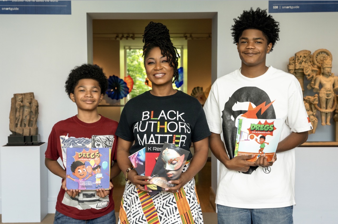 Three people, two children and one adult woman, stand indoors holding books, smiling, with artwork on walls behind them.