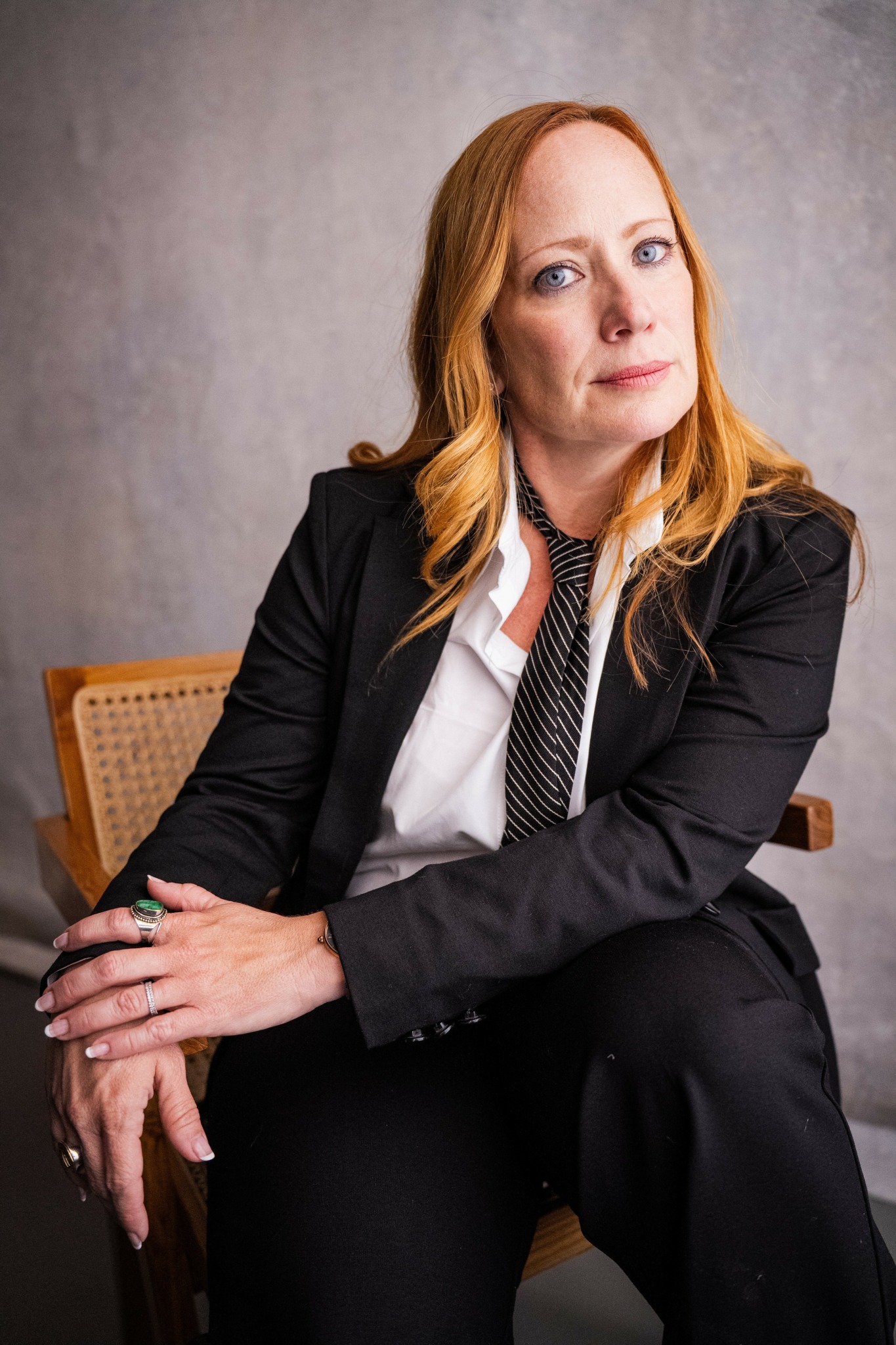 Woman with red hair in a black blazer and white shirt, sitting on a chair against a gray background.