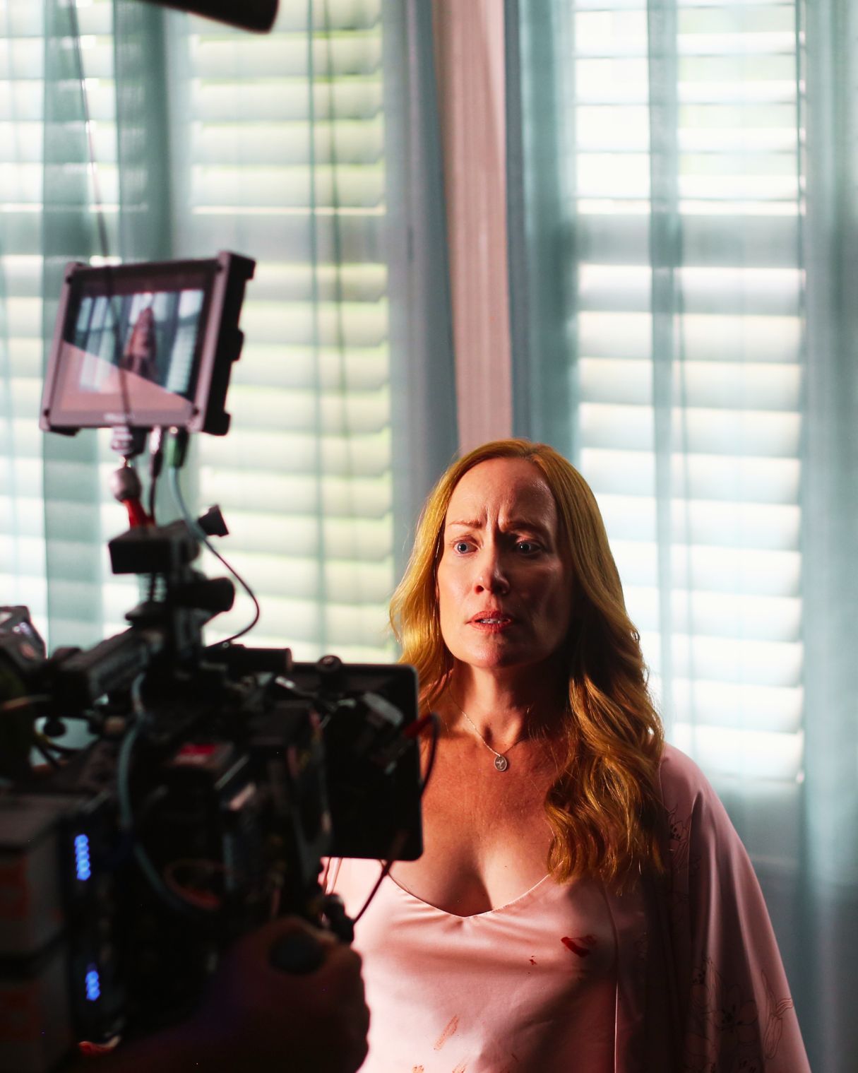 Woman with long hair in front of camera, illuminated by window light, appears to be speaking or reacting.