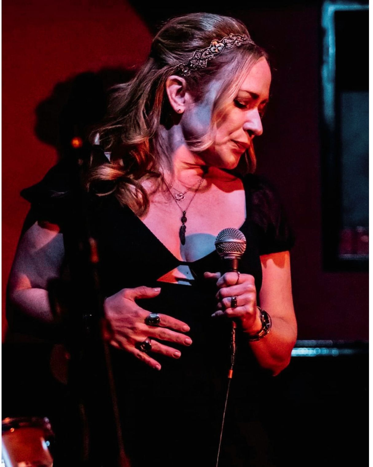 Woman holding a microphone, looking down, with curly hair and a black dress, on stage with red and blue lighting.