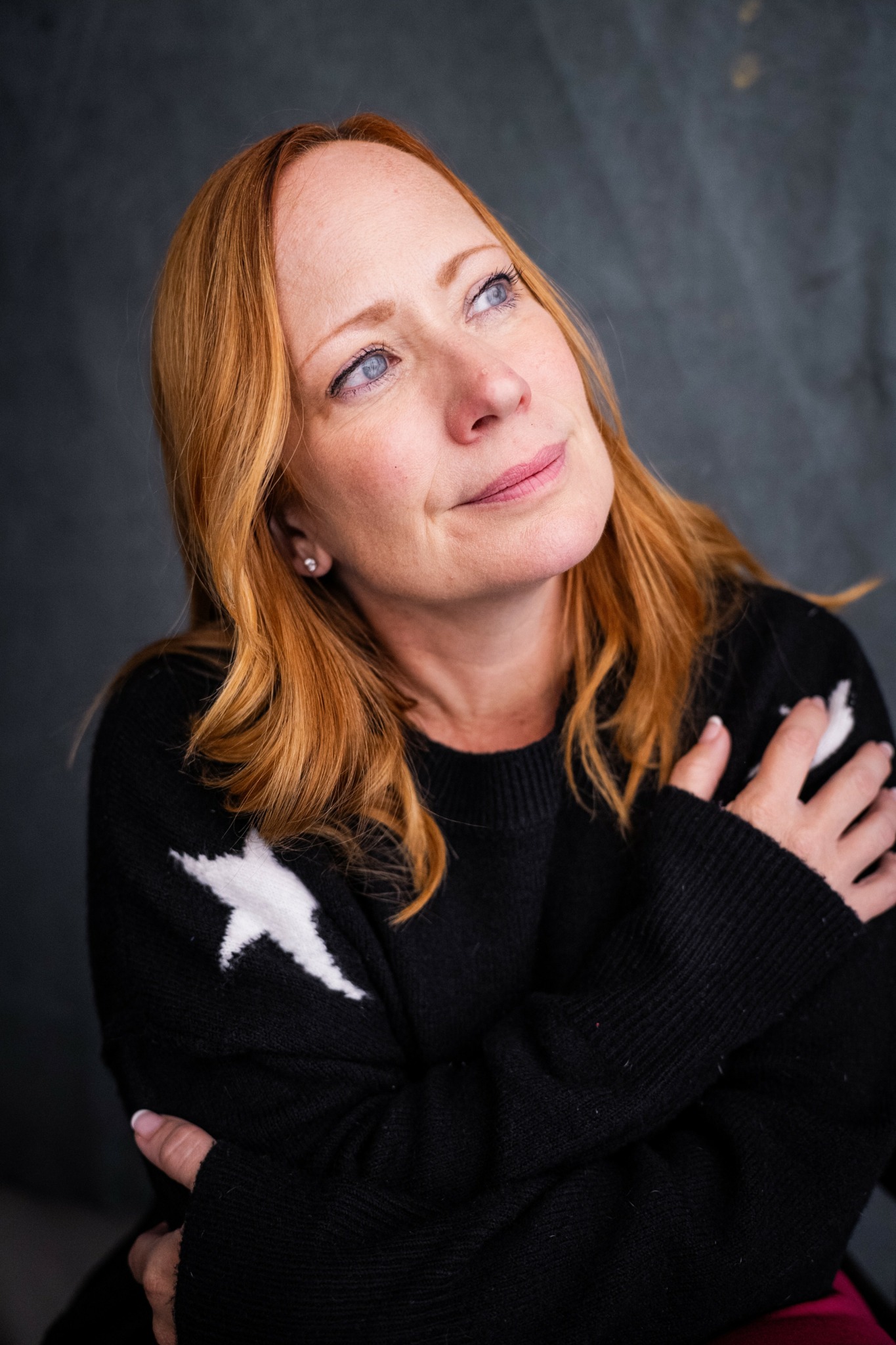 Woman with red hair looking upward, arms crossed, wearing a black sweater with white star pattern, against dark background.