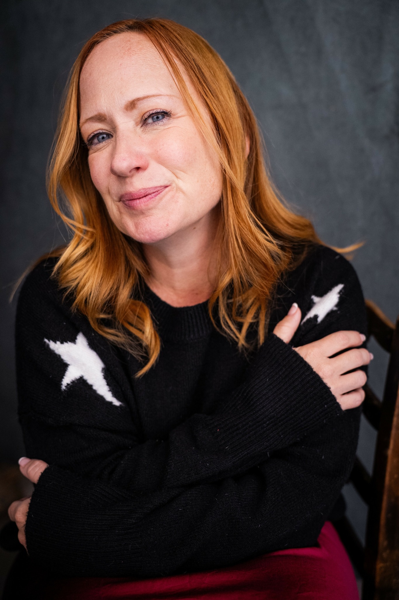 Woman with red hair smiling, wearing a black sweater with white star patterns, sitting with arms crossed against a dark background.