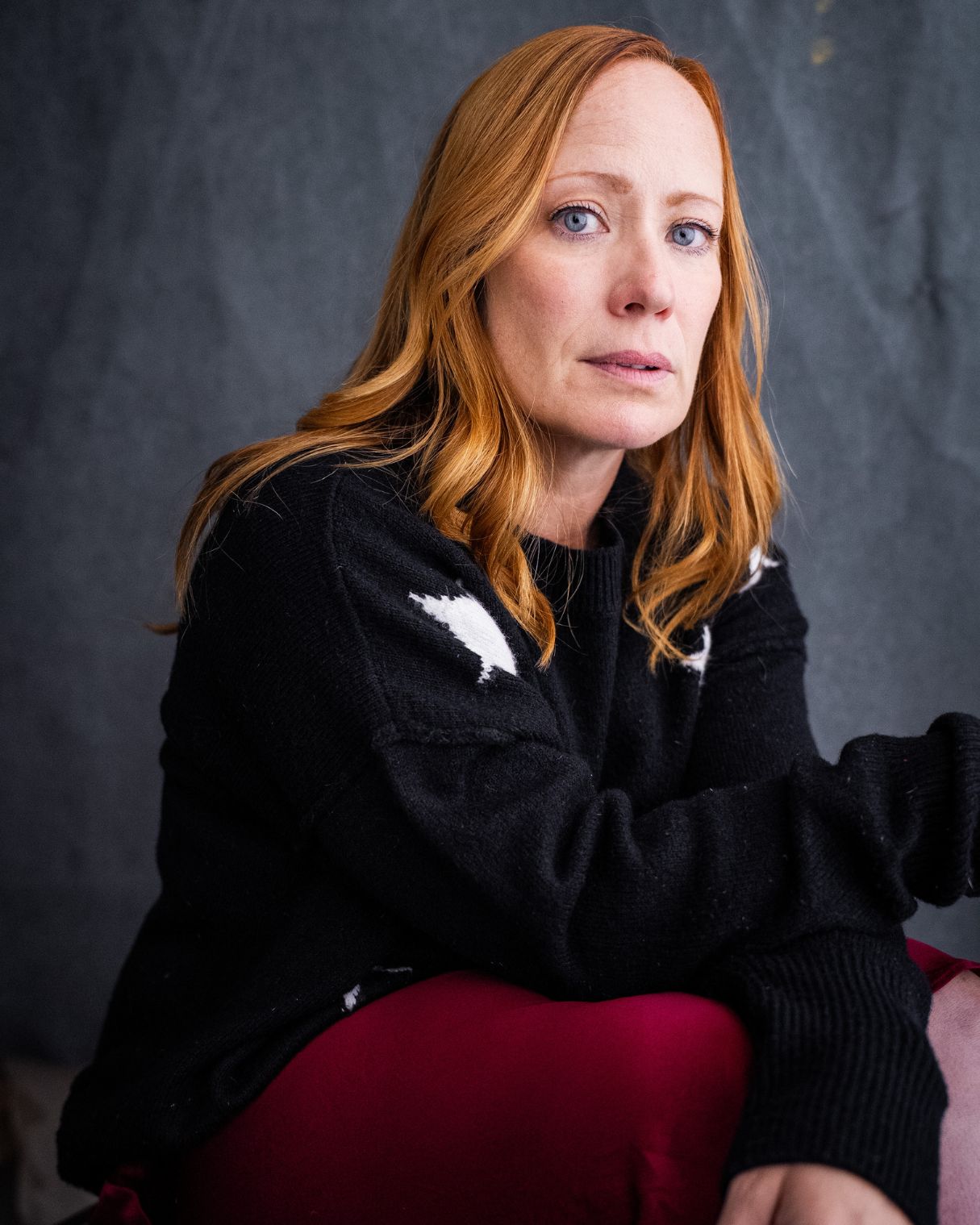 Woman with red hair wearing a black sweater, sitting against a dark background.