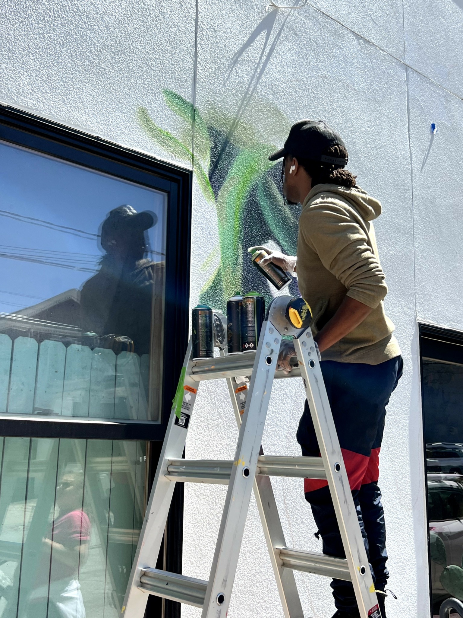 Person on ladder painting a mural on a wall, with cans of paint nearby, reflected in window, outdoors.