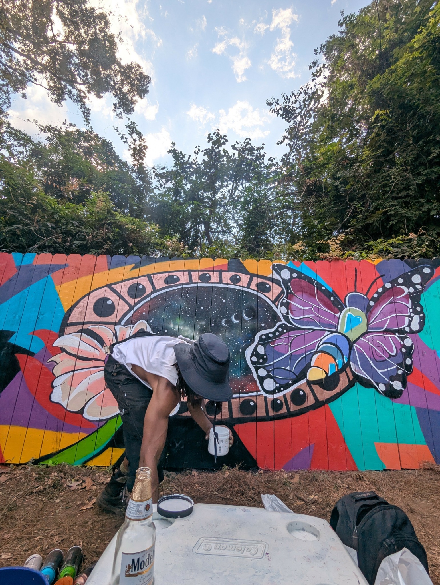 Person painting a colorful butterfly mural on a wall outdoors, surrounded by trees and sky, wearing a hat and white shirt.