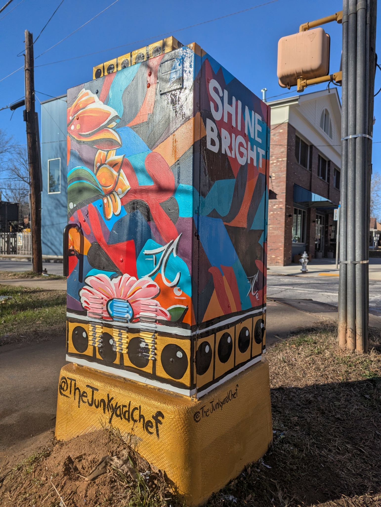 Colorful mural on a utility box with the words 'SHINE BRIGHT' and abstract shapes, located on a city sidewalk.