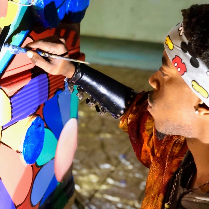 Person painting a colorful mural on a wall, wearing a patterned headband and a brown shirt, outdoors.