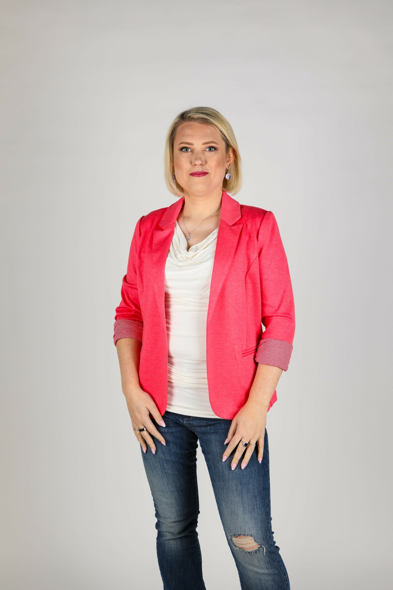 Woman with blonde hair wearing a pink blazer, white top, and jeans, standing against a plain background.