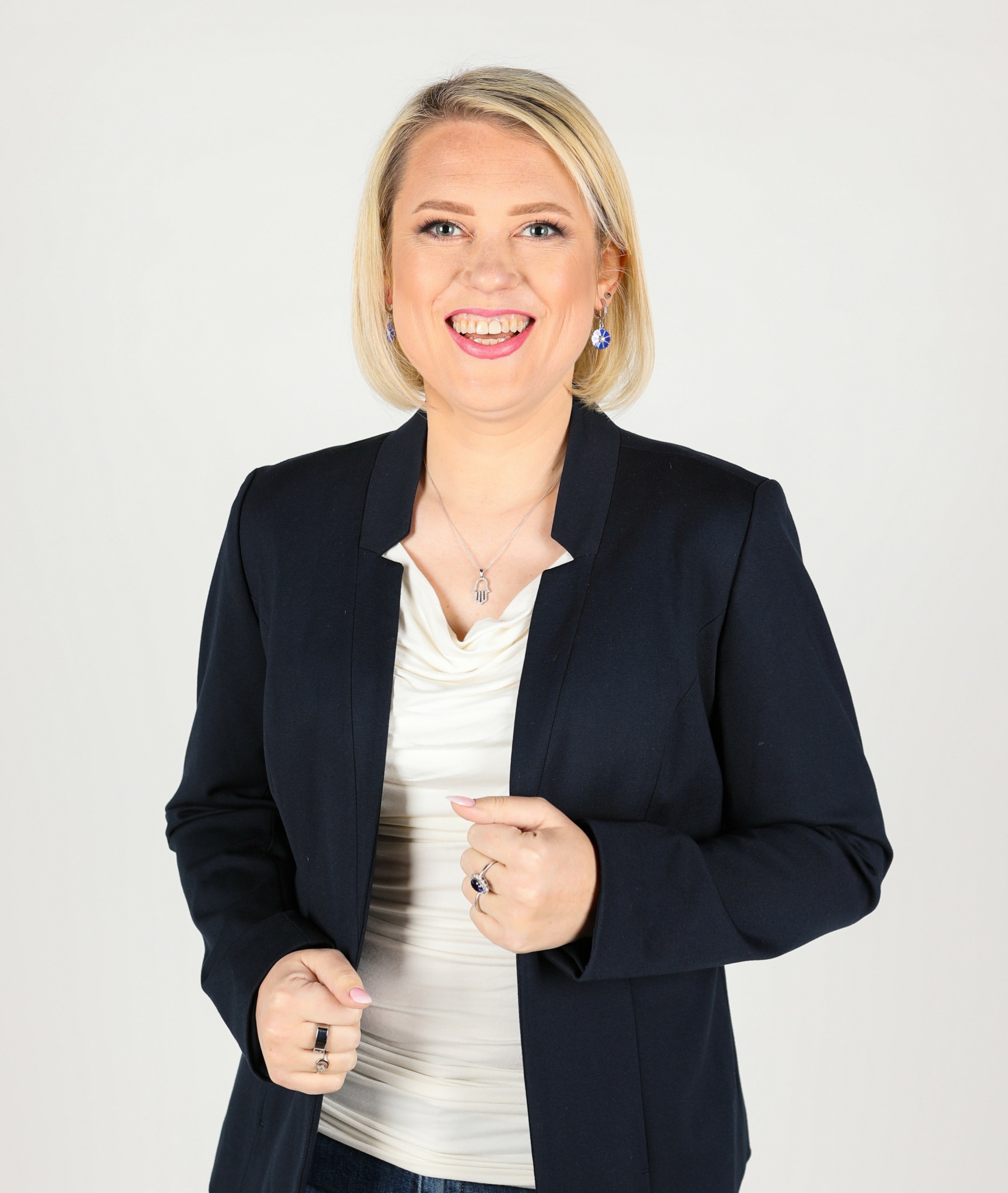 Smiling woman with blonde hair wearing a dark blazer and white top, standing against a plain background.