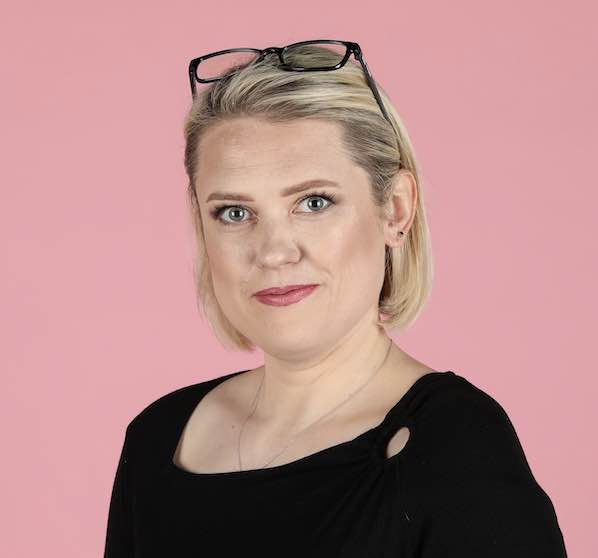 Woman with blonde hair, glasses on head, wearing black top, against pink background.