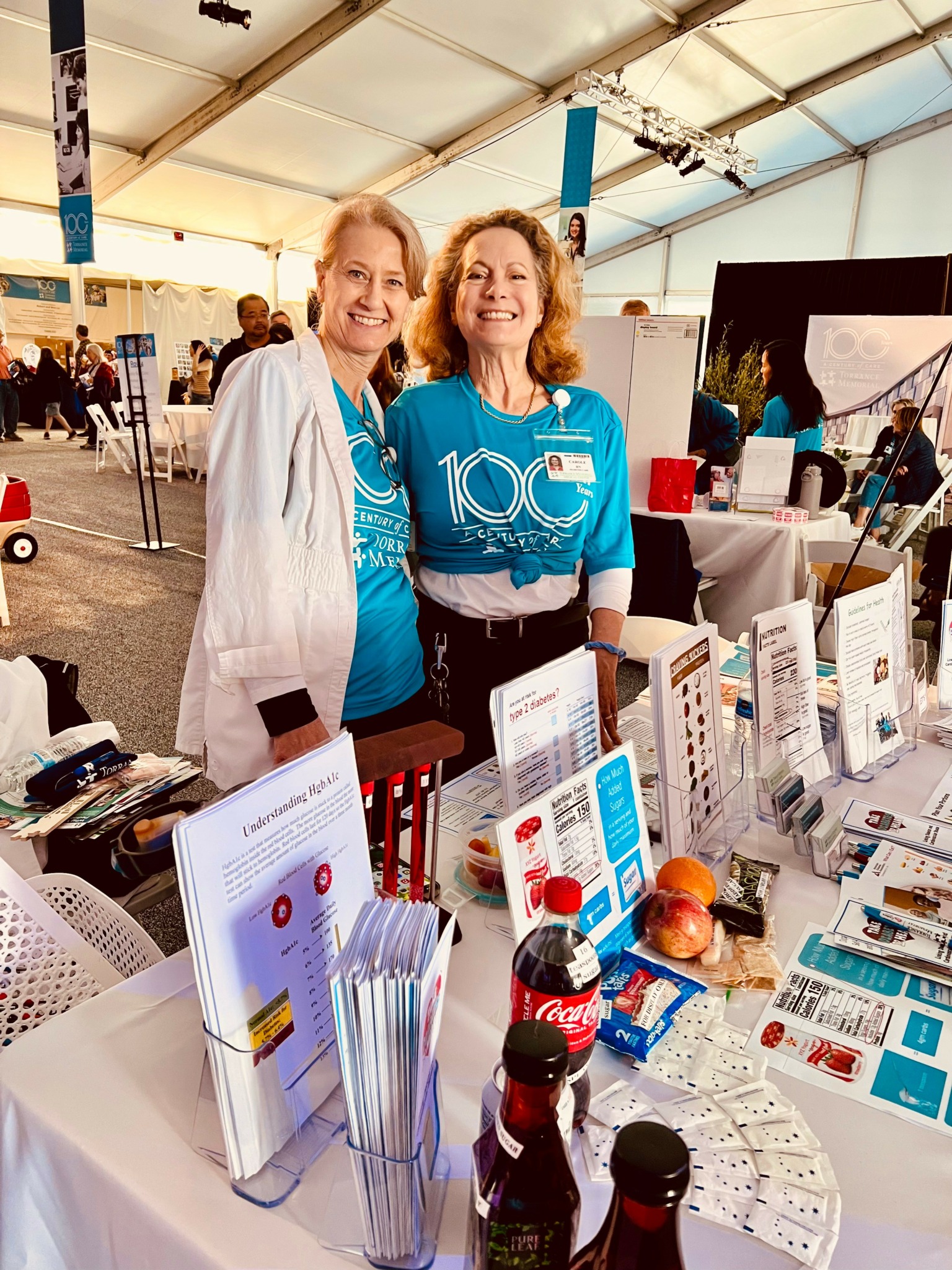 Two women smiling at a booth with informational displays and products inside a large tent, celebrating a 100th event or milestone.