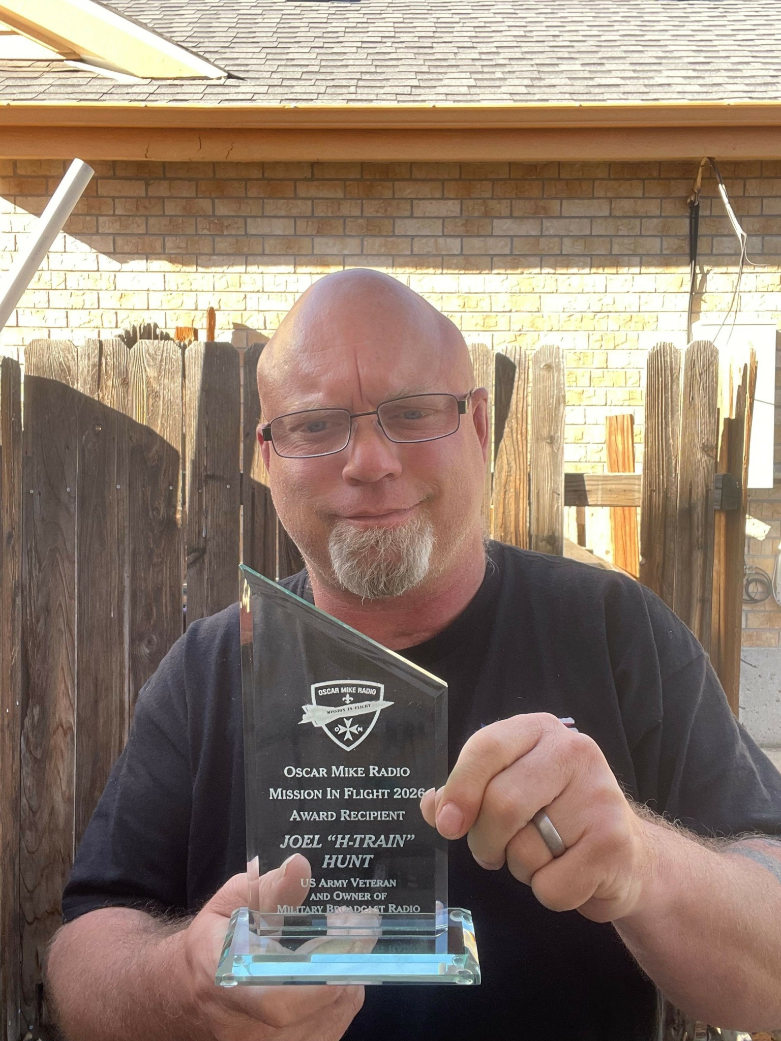 Man with glasses holding a glass award, smiling, in a room with brick walls and wooden beams.